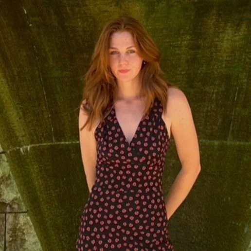 Red-haired woman in a black dress with red polka dots standing outdoors in front of a tree.
