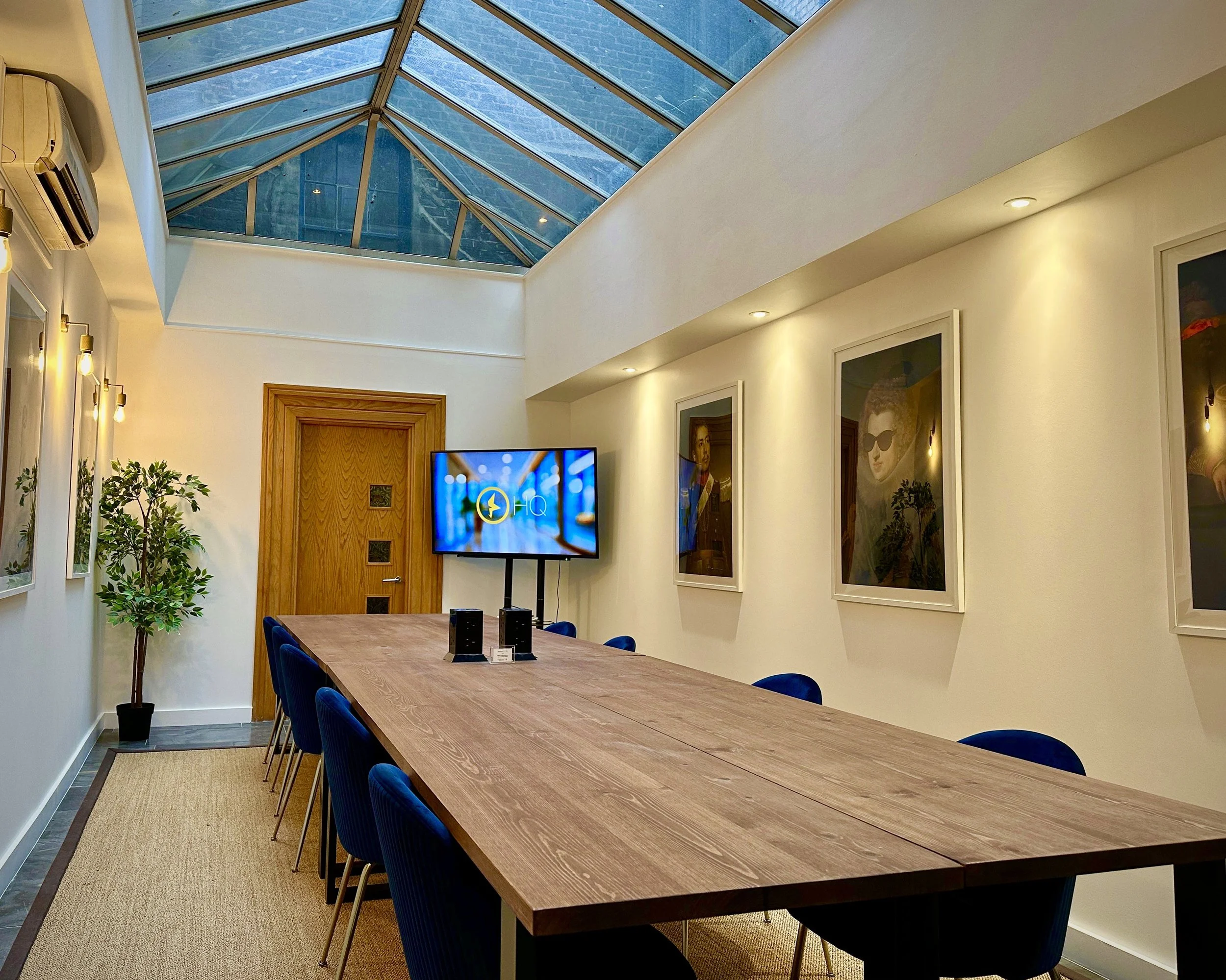 A picture of a light and airy meeting room with a long wooden table and blue velvet chairs with a big TV screen and a conservatory-style ceiling.