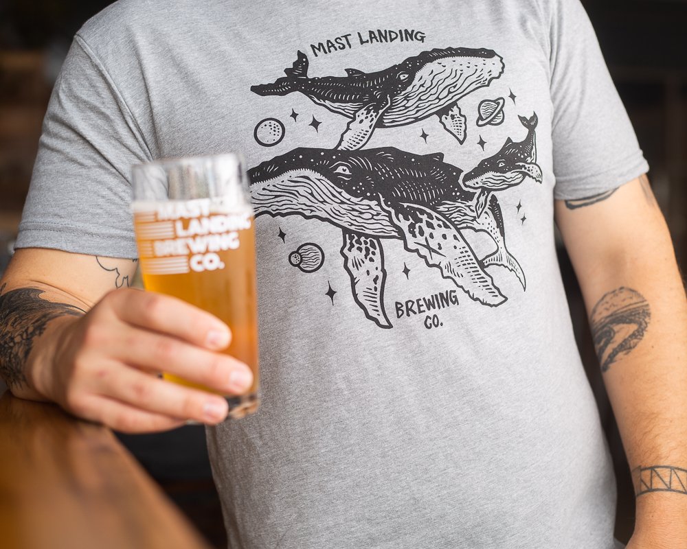 Mast Landing’s “Space Whales” tee sports three whales swimming amongst the planets in space. “Mast Landing Brewing Co.” in handwritten style is printed in black ink across the back neck of the shirt.
