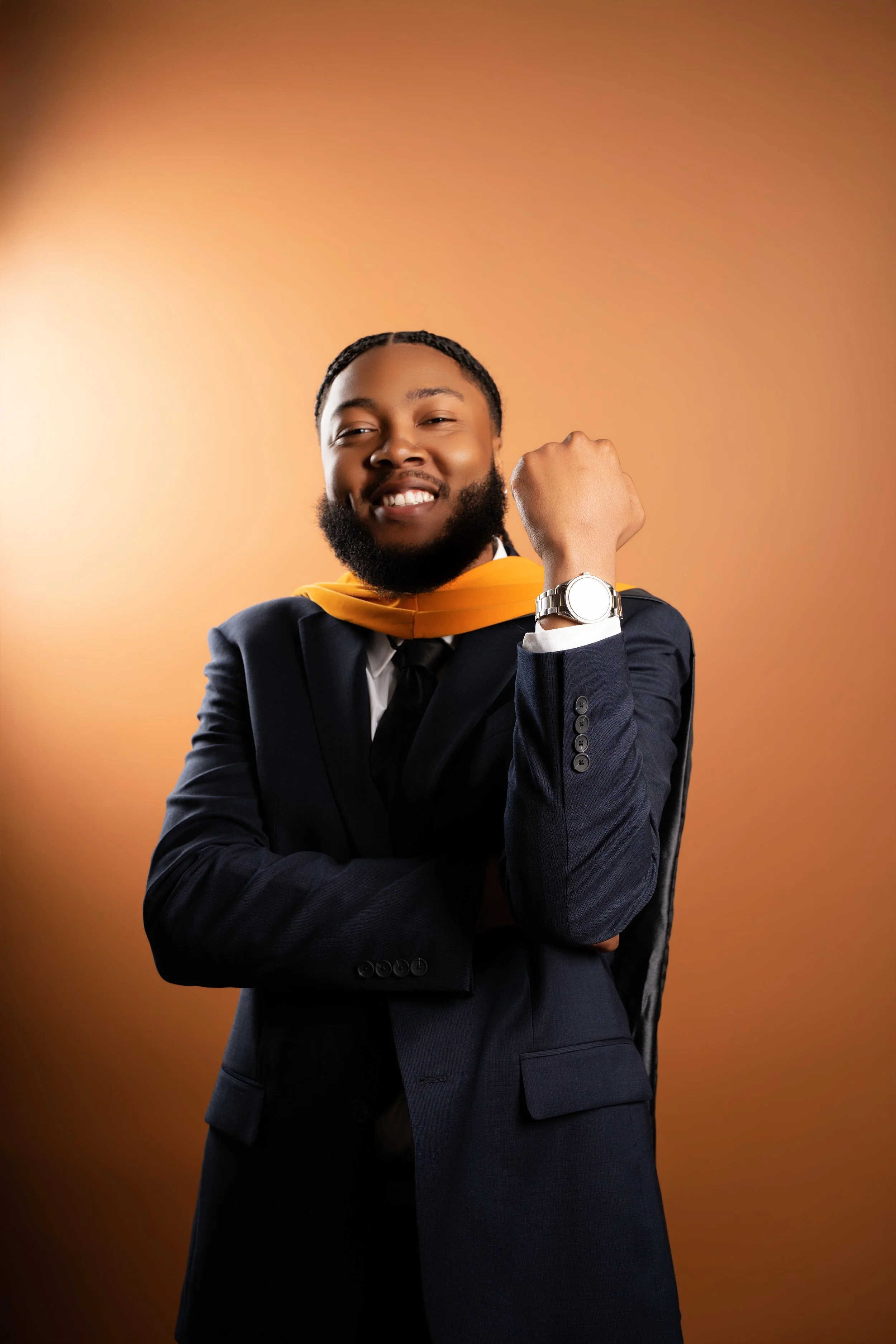 Young man with curly hair and beard in a business suit, wearing a yellow graduation stole and a silver wristwatch, smiling confidently against a warm orange background.