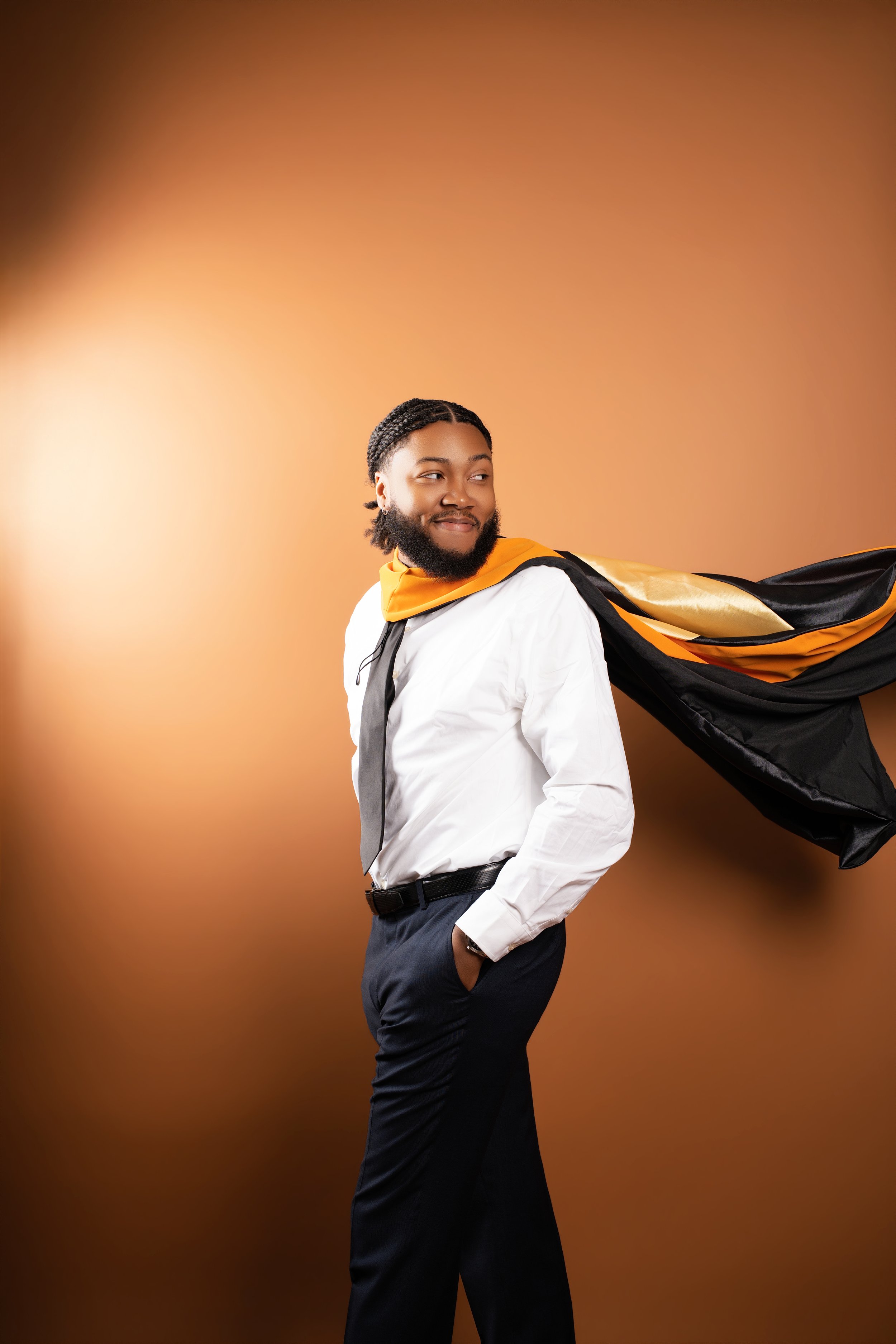 A man with braided hair and a beard, smiling slightly, wearing a white shirt, dark trousers, and a black belt. He has a yellow and black cape draped over his shoulder and is standing against a brown gradient background.