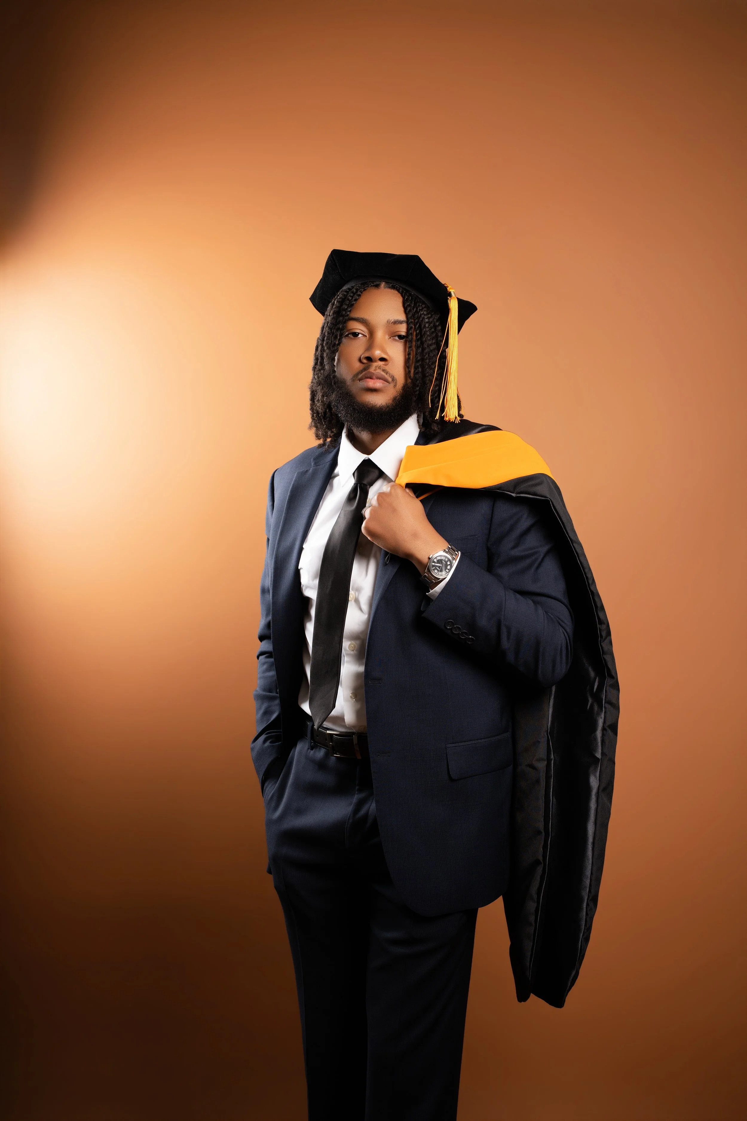 A man dressed in a dark suit, white shirt and black tie, holding a black graduation cap with yellow tassels over his shoulder, standing against a warm sepia-colored background.