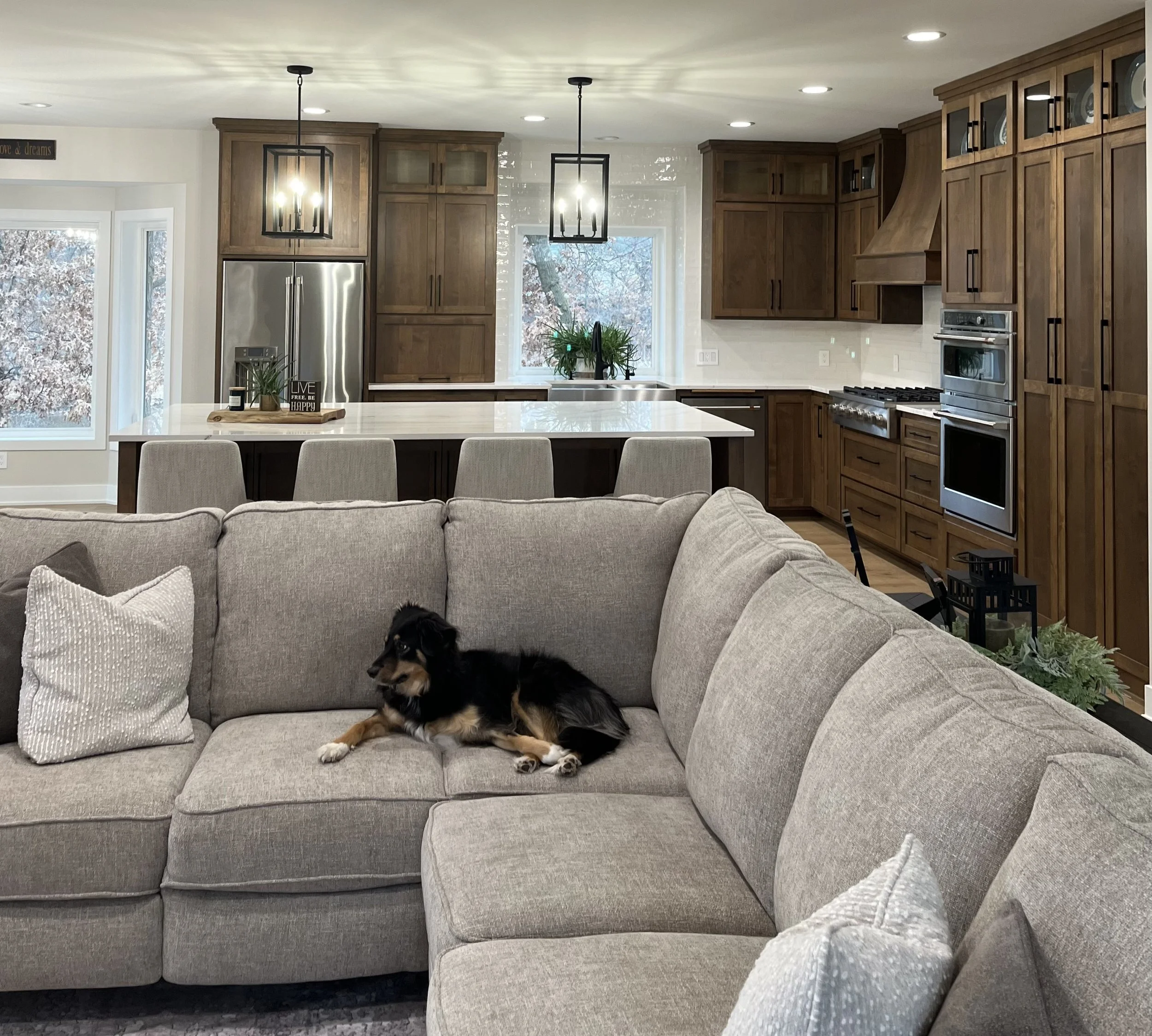 Soft neutral sectional sofa with warm wood custom cabinetry in the kitchen space behind the living room. Stainless cafe appliances, white quartz countertops, white subway tile, and modern geometric pendant lighting