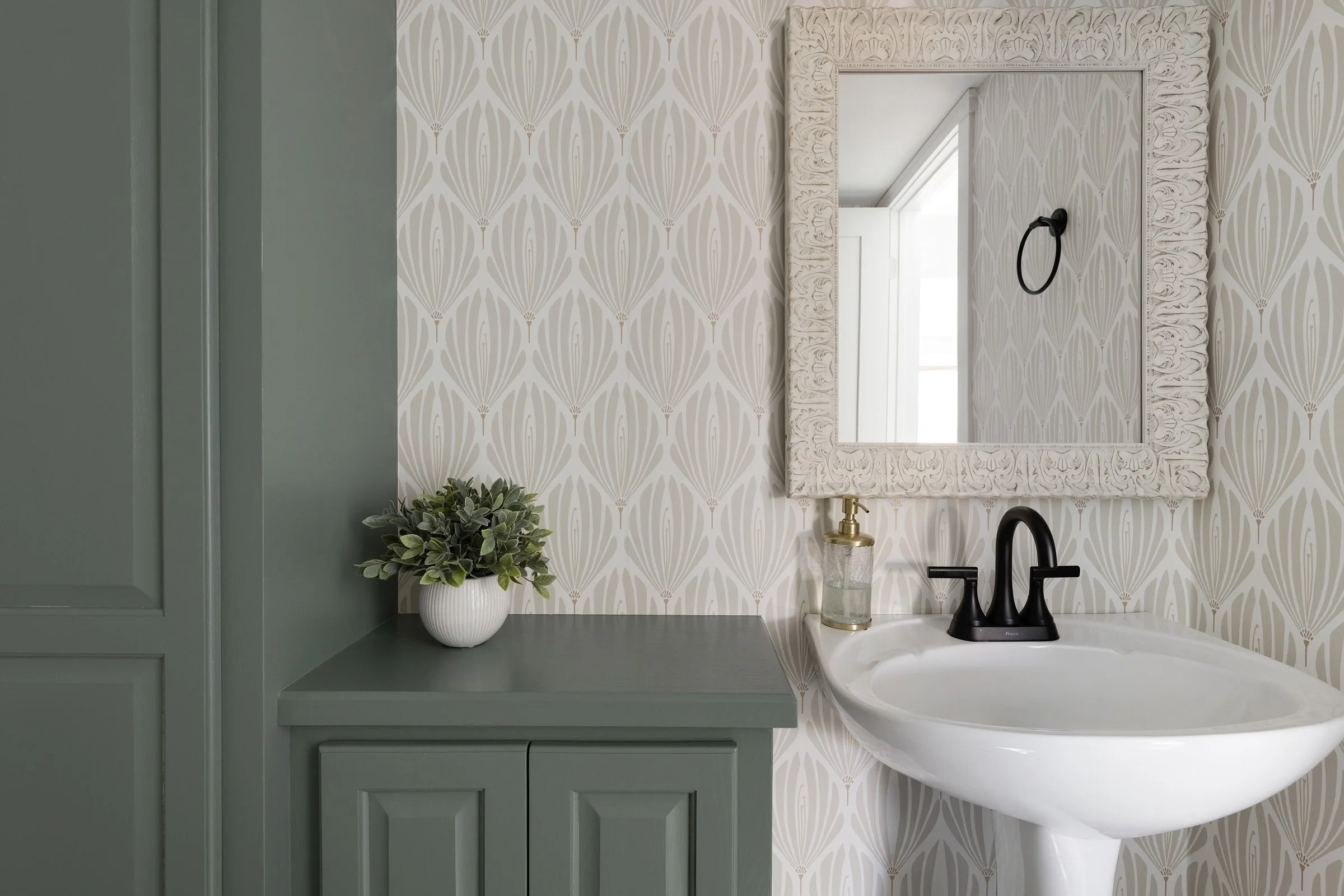 A bathroom vanity area with a green cabinet, a small potted plant, a soap dispenser, a black faucet, and a large framed mirror on a patterned wallpapered wall.