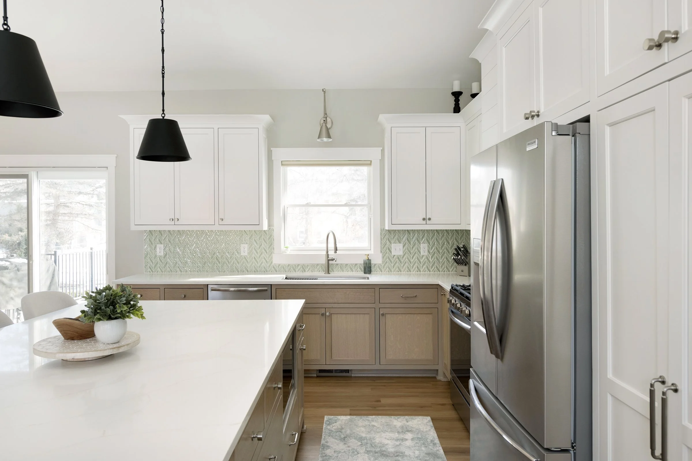 Bright kitchen with white upper cabinets, beige lower cabinets, a stainless steel refrigerator, a large white countertop island, hardwood flooring, pendant lights, a window over the sink, and a green patterned backsplash.