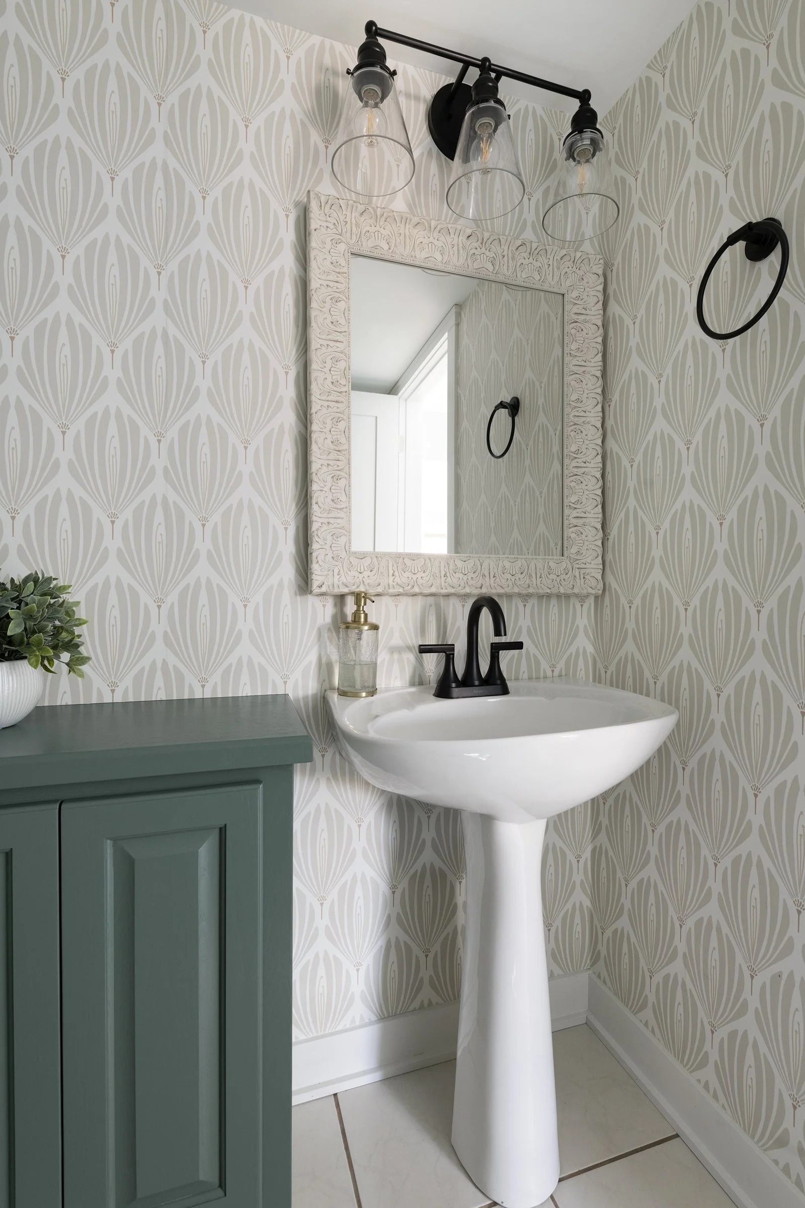 A small bathroom with a white pedestal sink, a framed mirror, a black faucet, a green cabinet, wallpaper with a neutral geometric pattern, a wall-mounted black towel ring, a modern black light fixture with three bulbs, and a soap dispenser on the sink.