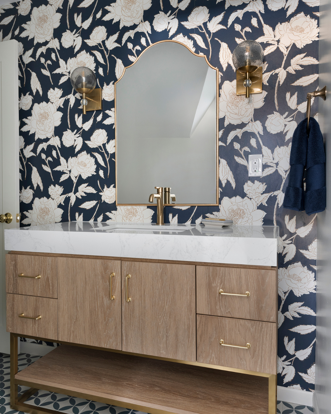 Bathroom vanity with a white marble countertop, gold hardware, and a wooden cabinet. A mirror with a gold frame, wall sconces, a dark floral wallpaper, and blue towels hanging on a gold hook.