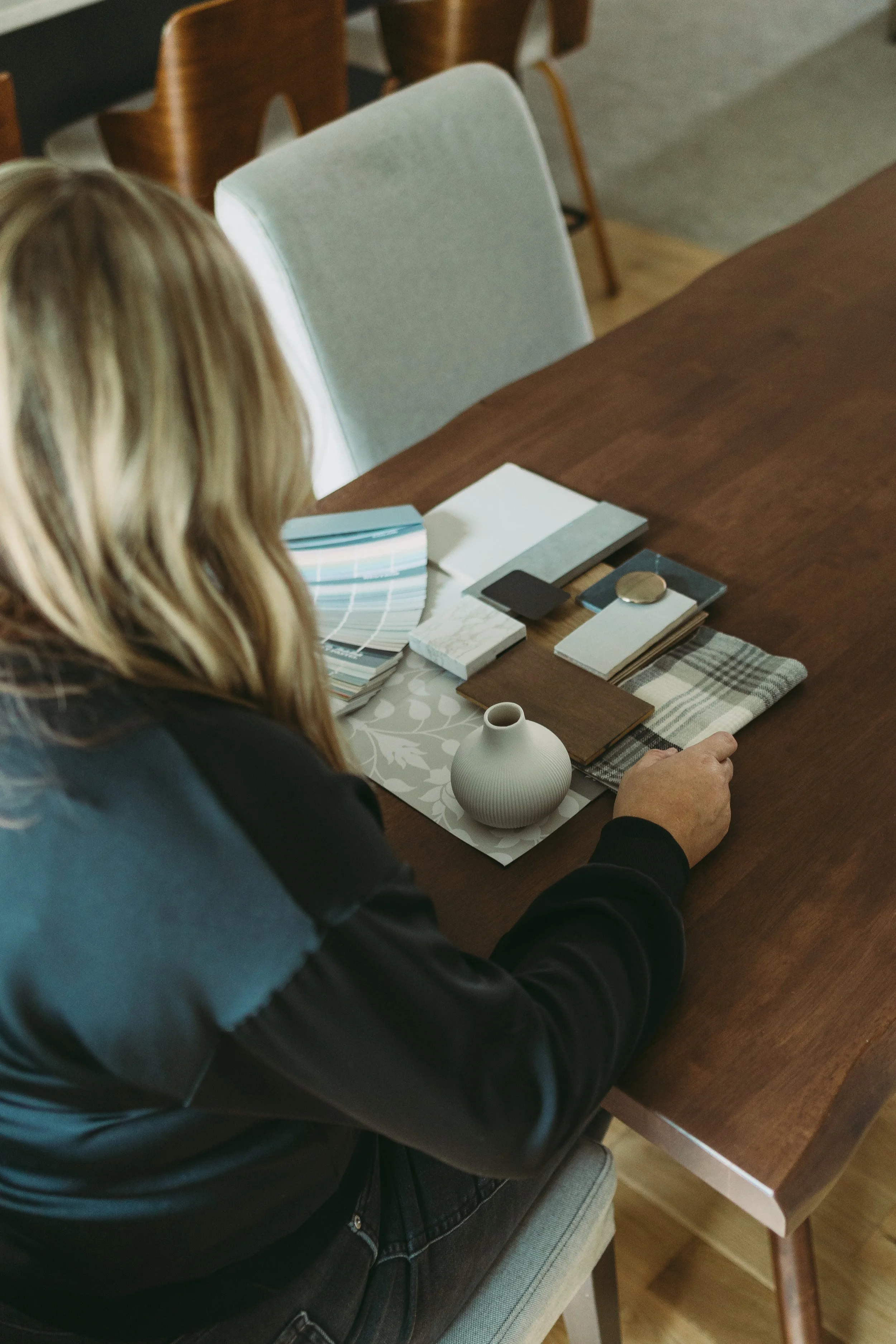 Woman sitting at a wooden table with a color palette, fabric swatches, color sample cards, and a small decorative vase.