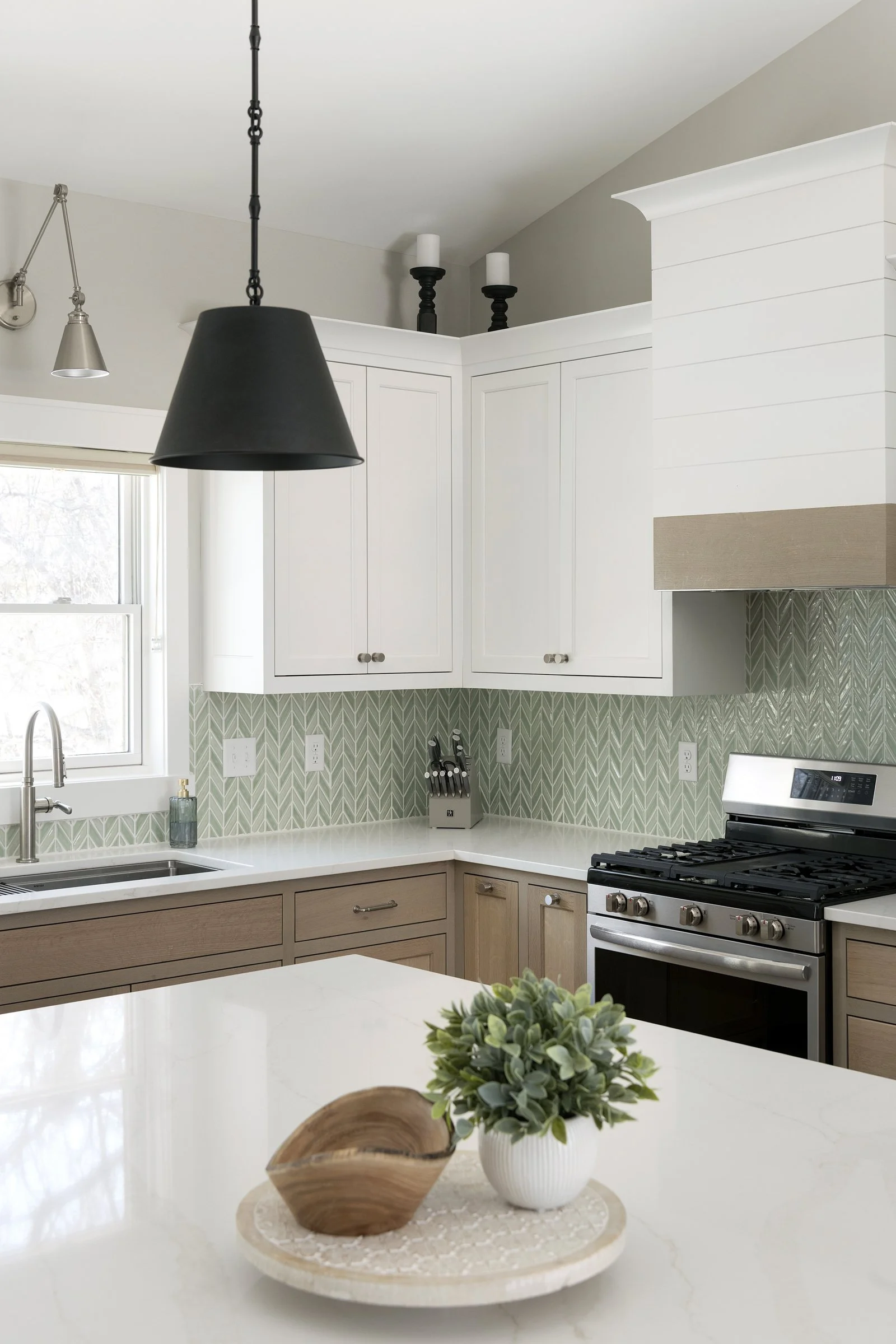 Modern kitchen with white upper cabinets, beige lower cabinets, a black pendant light, a window above the sink, patterned green tile backsplash, a stainless steel gas stove, and a decorative potted plant and wooden bowl on the white marble countertop.
