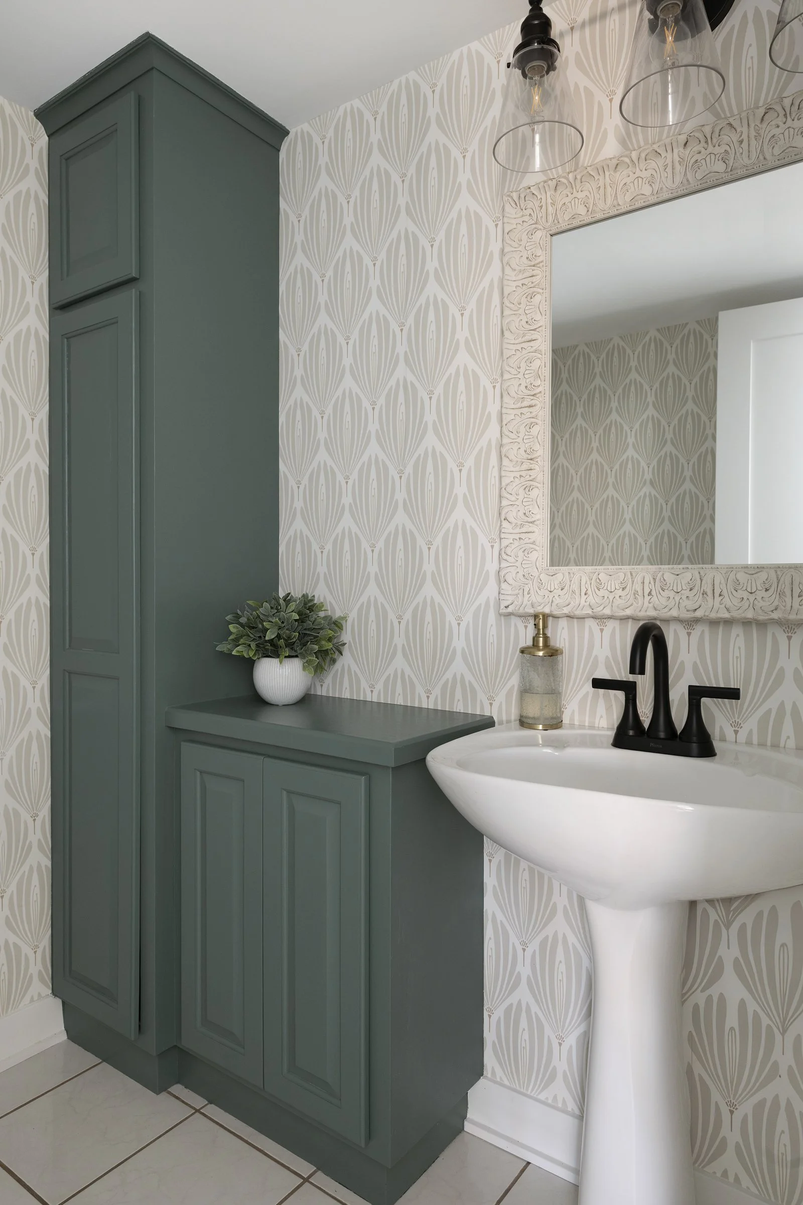 Bathroom with patterned wallpaper, green cabinet, white sink, black faucet, mirror, and a small plant.
