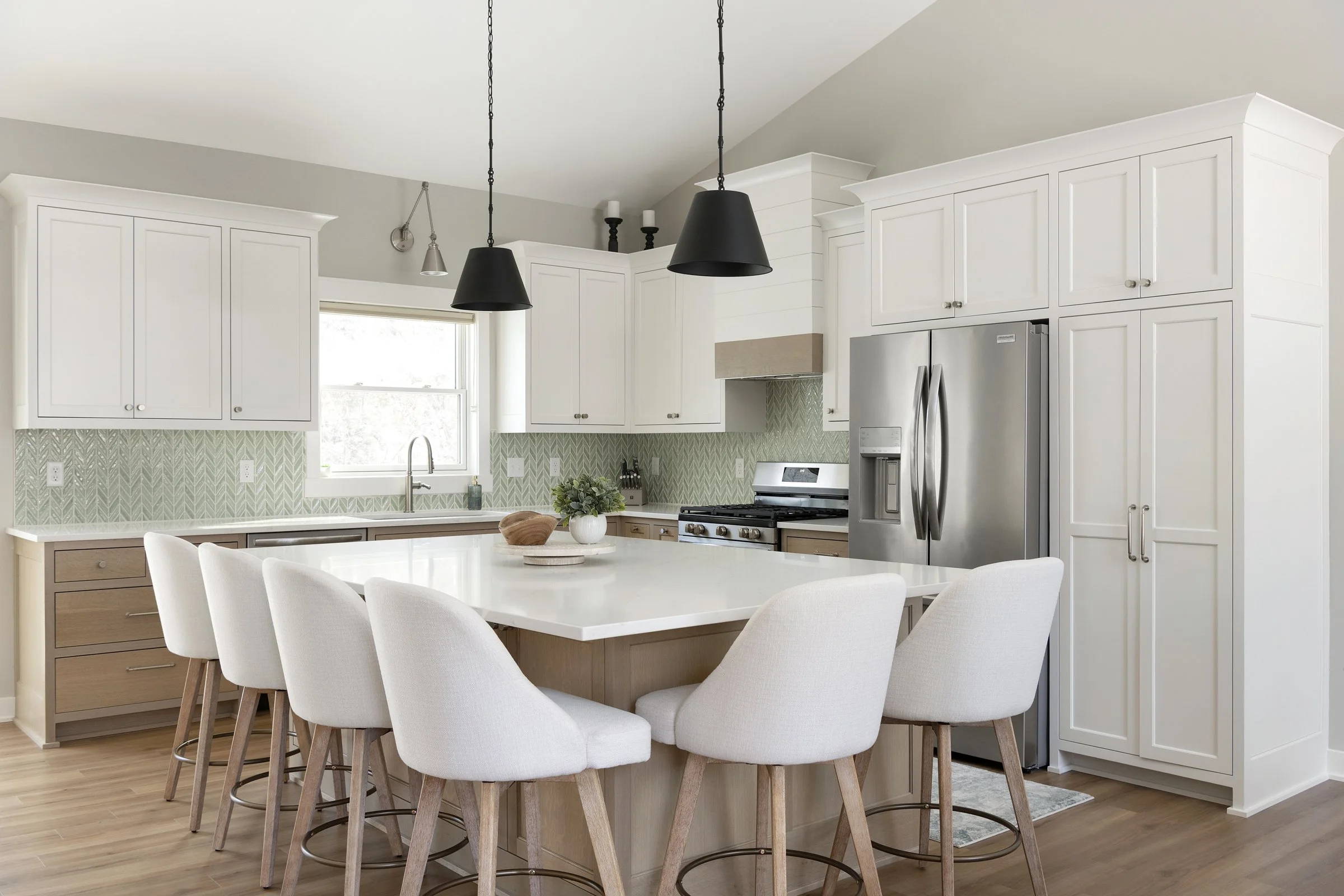 Modern kitchen with white cabinetry, stainless steel refrigerator, island with seating, black pendant lights, and a window above the sink.