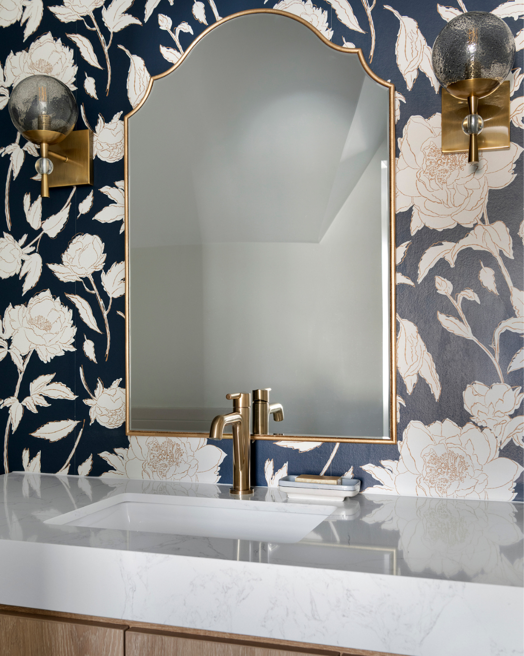 A bathroom sink with a marble countertop, a gold faucet, a rectangular tray, a large mirror with a gold frame, floral patterned wallpaper, and two wall sconces with cloudy glass globes and gold accents.