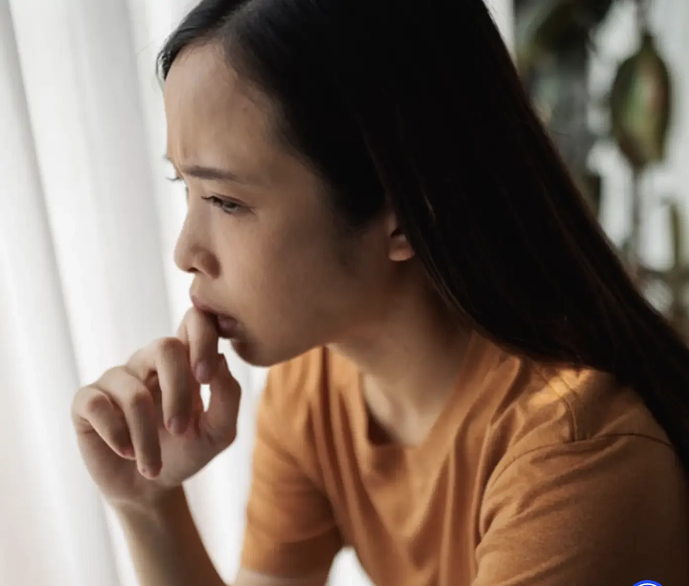 A woman with dark hair and light skin, wearing a yellow shirt, is sitting near a window with white curtains, appearing to be deep in thought or worried.