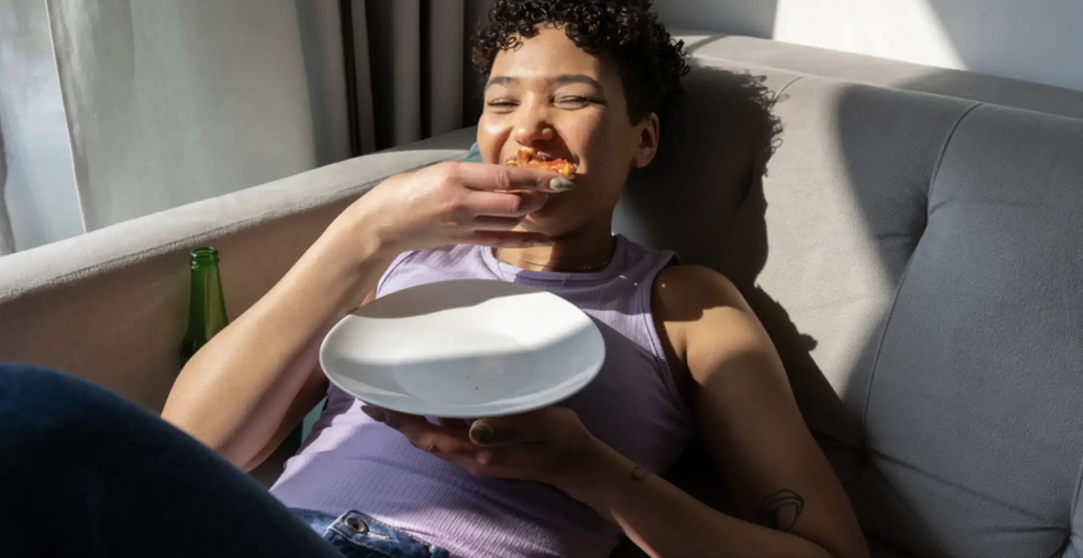 Young woman with short curly hair laughing and eating pizza while relaxing on a beige couch at home, with a green bottle nearby.