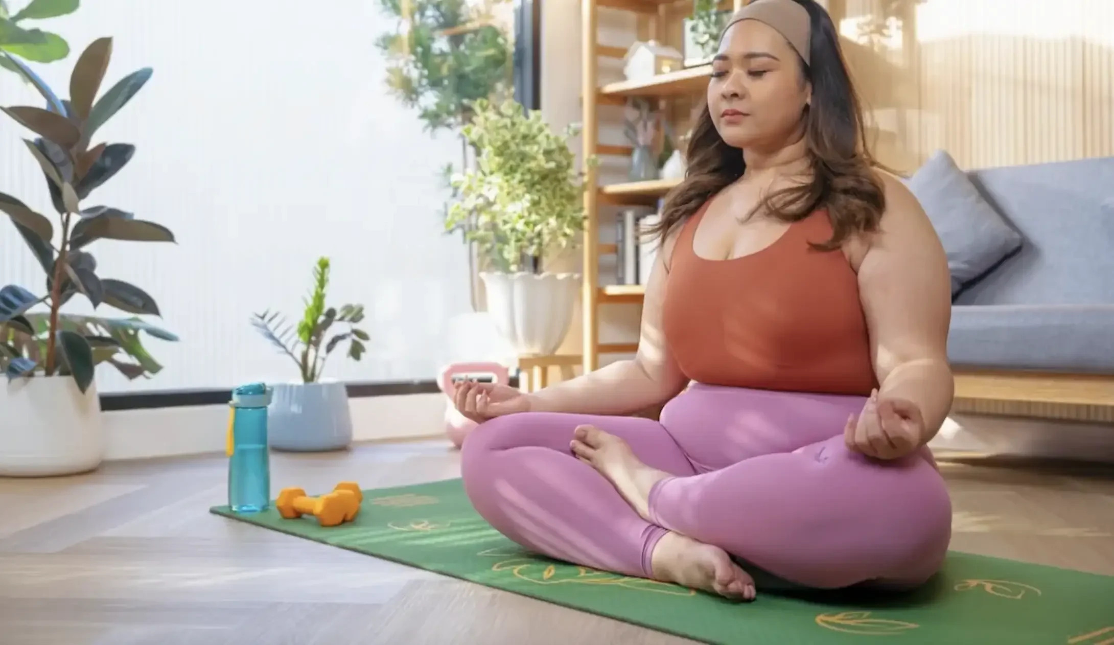 A woman practicing yoga indoors, sitting cross-legged on a green yoga mat with a water bottle, orange dumbbells, and meditation beads nearby. She has long dark hair, a headband, and is wearing a brown tank top and pink leggings.
