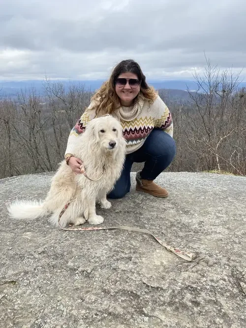 A woman with curly hair wearing sunglasses, a beige sweater with colorful patterns, and jeans, is crouching on a large rock with a cream-colored, long-haired dog on a leash. They are outdoors in a mountainous area with leafless trees and a cloudy sky in the background.