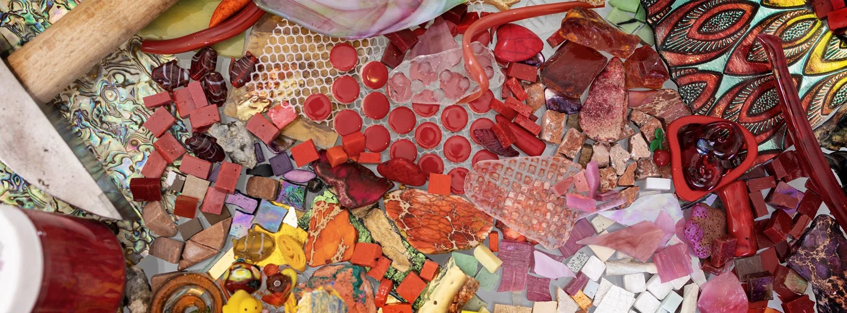 Colorful mosaic materials including various shapes and colors of glass, tiles, and stones, arranged on a surface. A hammer is visible on the left side.