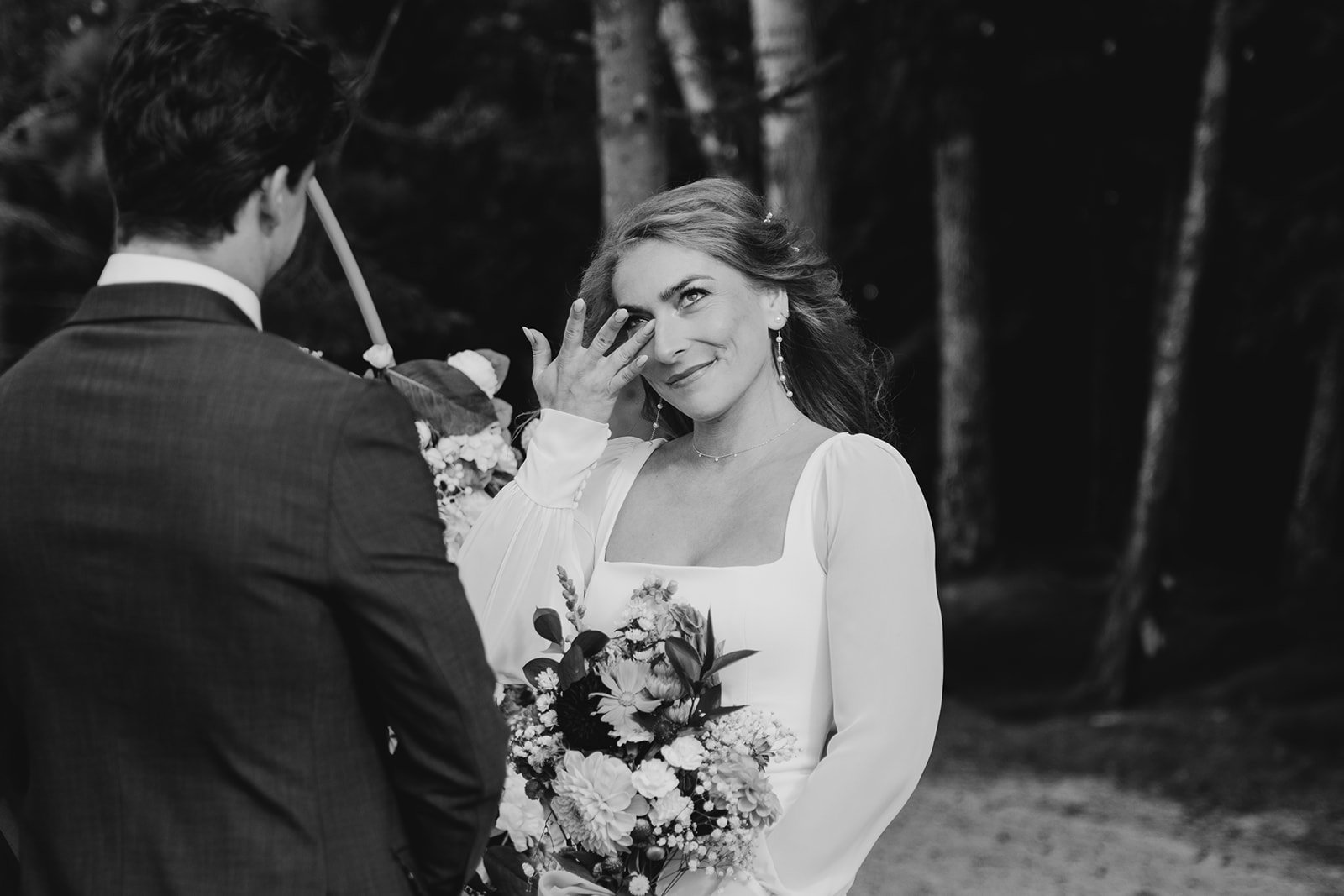 A woman in a white dress holding a bouquet of flowers, smiling as she wipes away a tear during a wedding ceremony outdoors, with a man in a suit partially visible facing the woman, trees in the background.