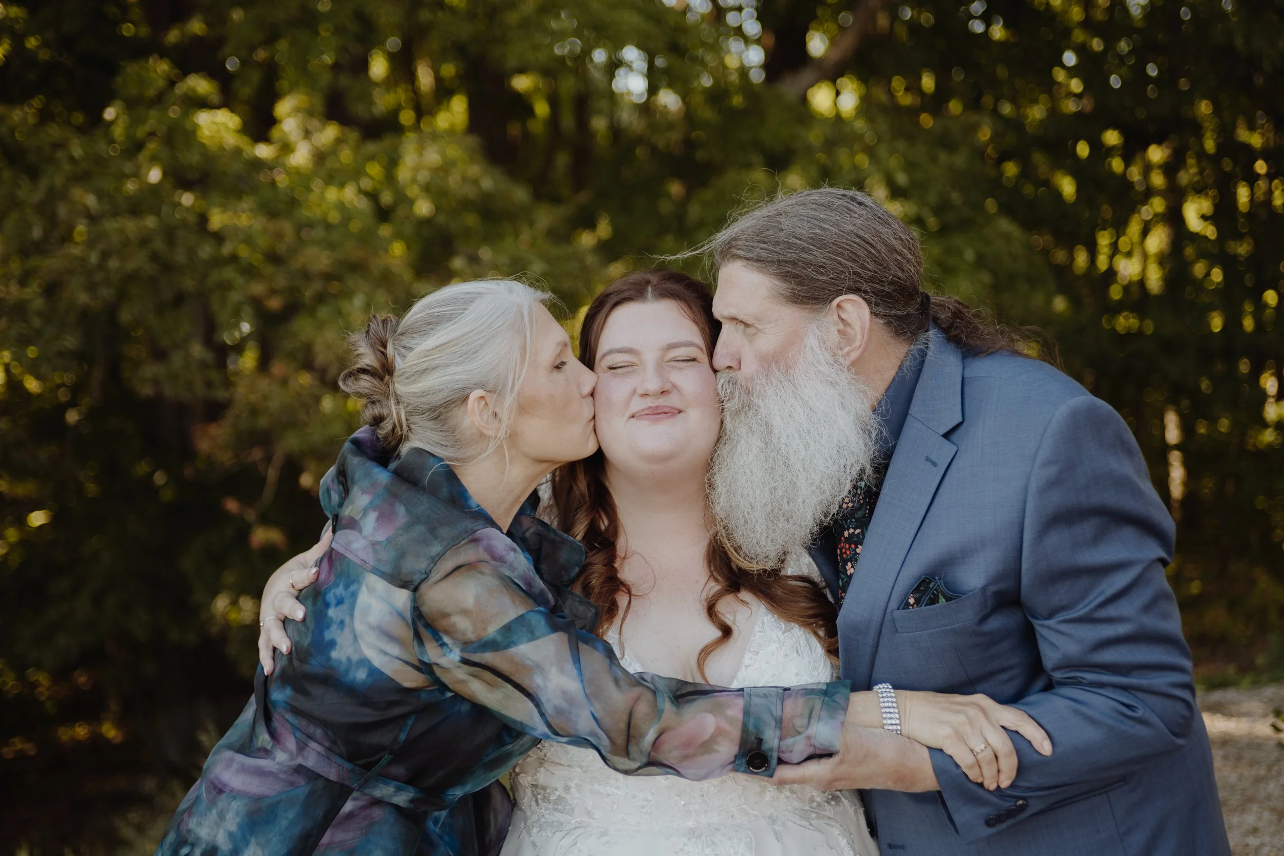 A woman in a wedding dress is kissed on the cheeks by an older woman with gray hair and an older man with a beard, outdoors with trees in the background.