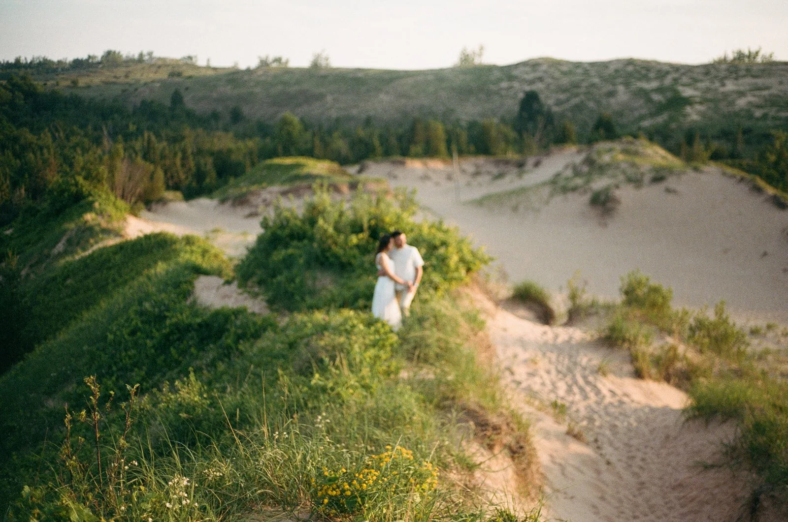 A couple stands on sandy dunes surrounded by green shrubs and wildflowers, with hills and forest in the background during sunset.