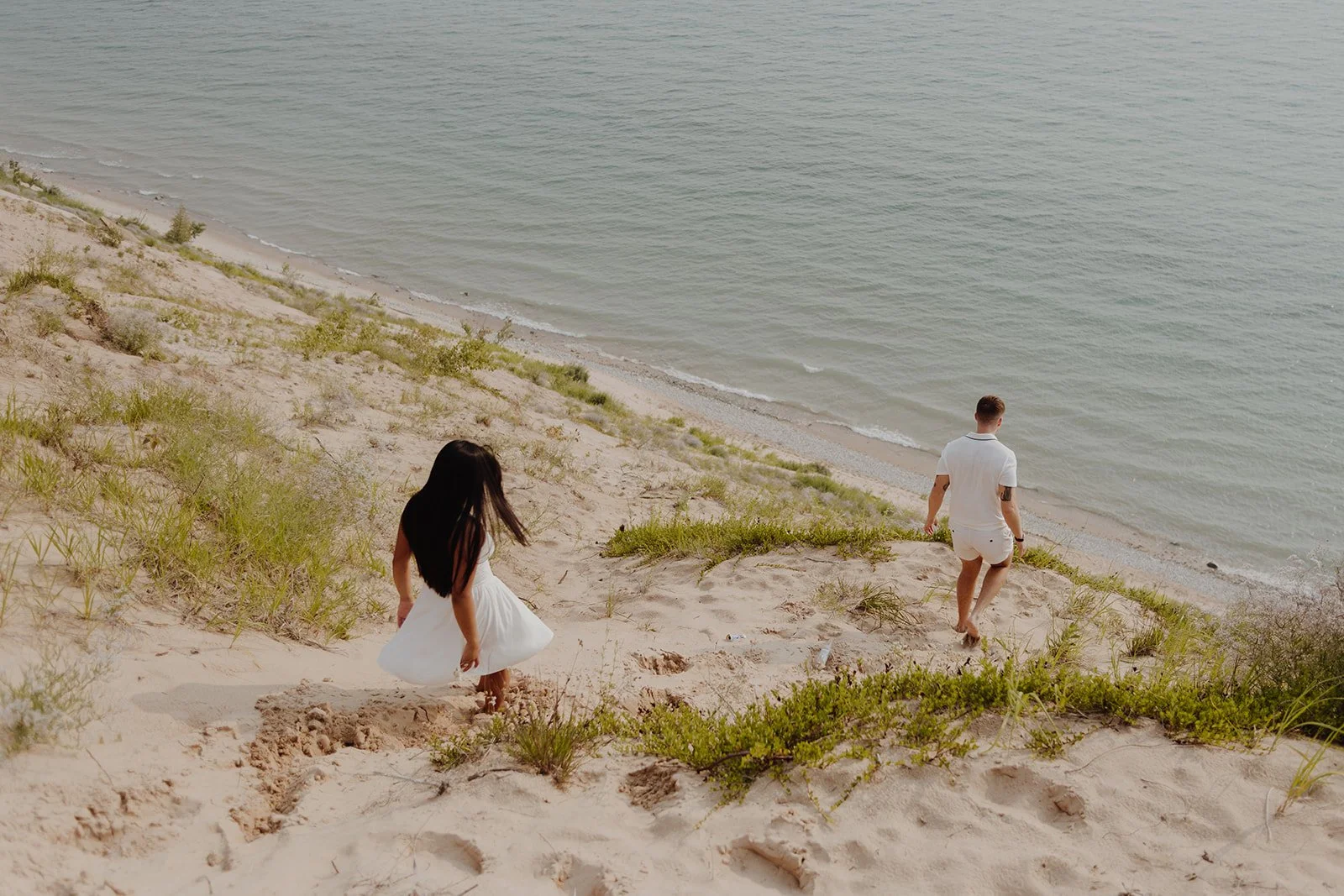 A woman in a white dress and a man in white clothes walking down a sandy hill toward a calm beach.