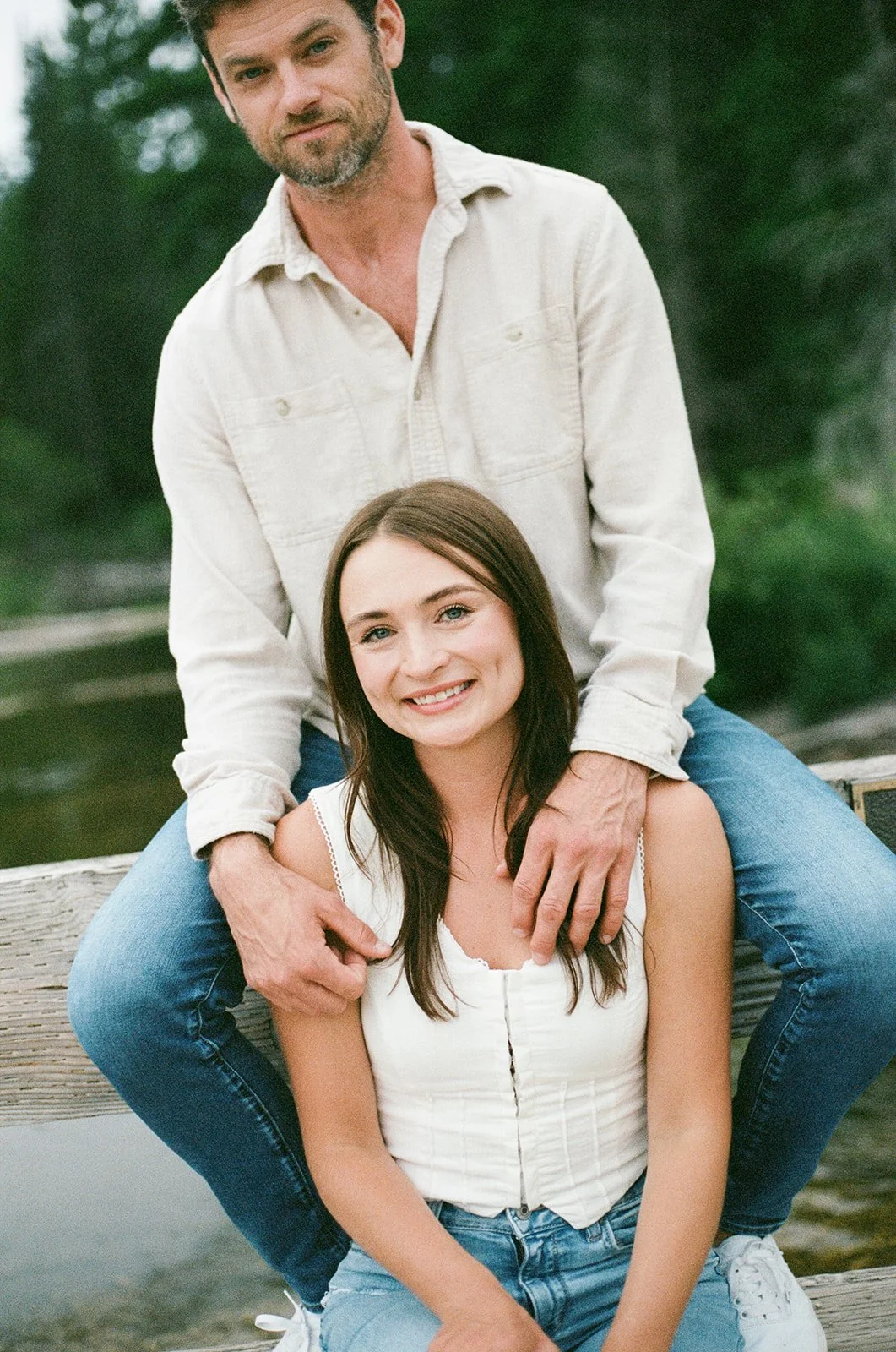 A young woman with long dark hair smiling while sitting on a wooden dock, with a man with a beard and short hair standing behind her, resting his hands on her shoulders. They are outdoors near water, with trees in the background.