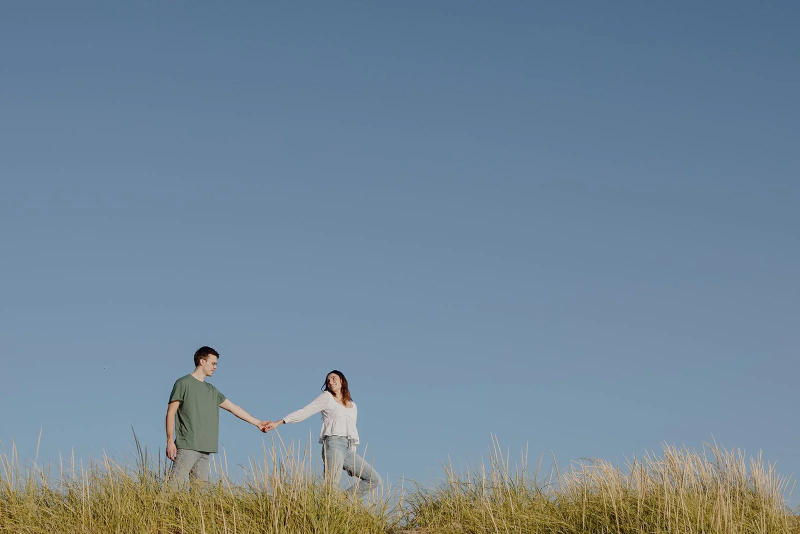 A young man and woman holding hands in a grassy field under a clear blue sky.