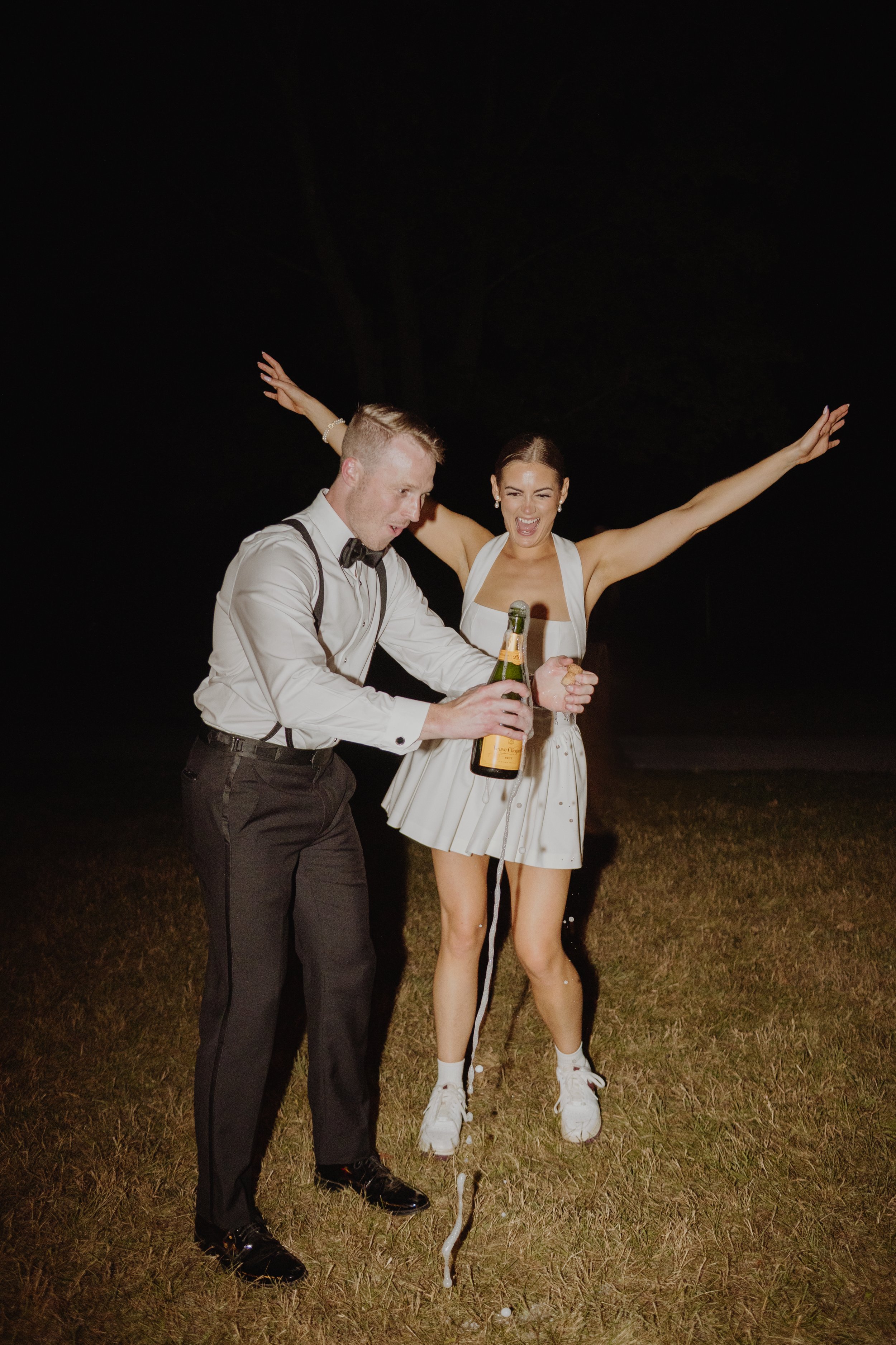 A man and a woman celebrating outdoors at night, with the man opening a champagne bottle and the woman smiling with arms raised.