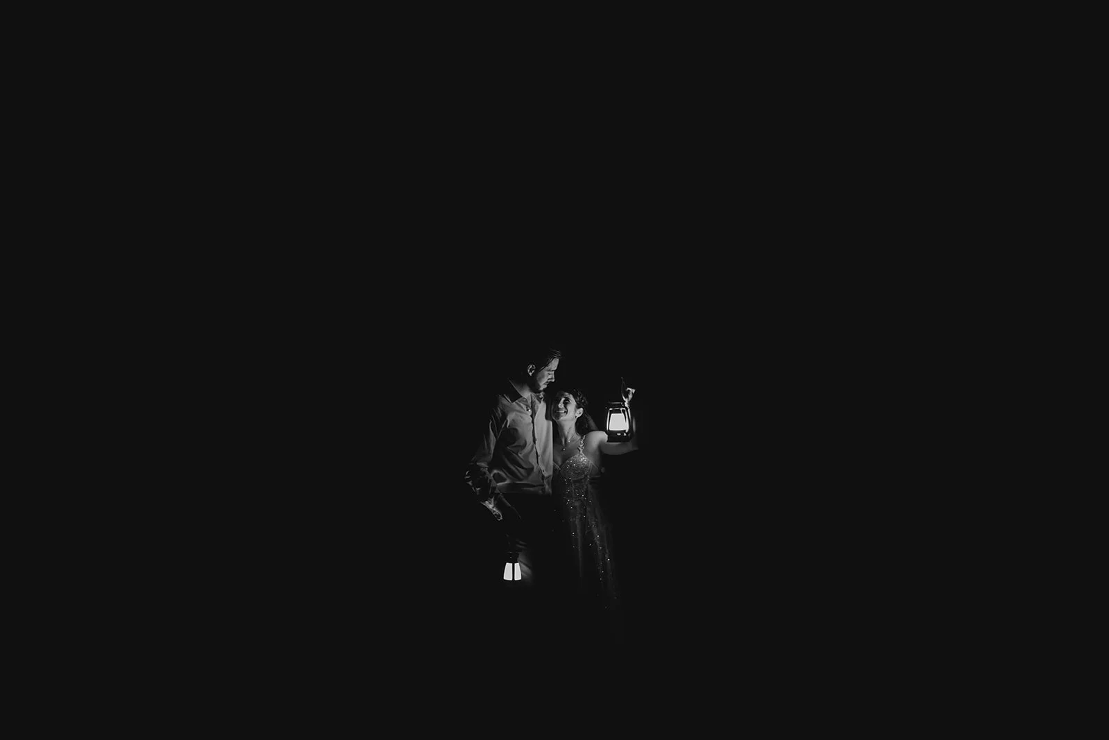 A woman and man in formal attire holding lanterns at night, with minimal lighting highlighting their faces and attire.