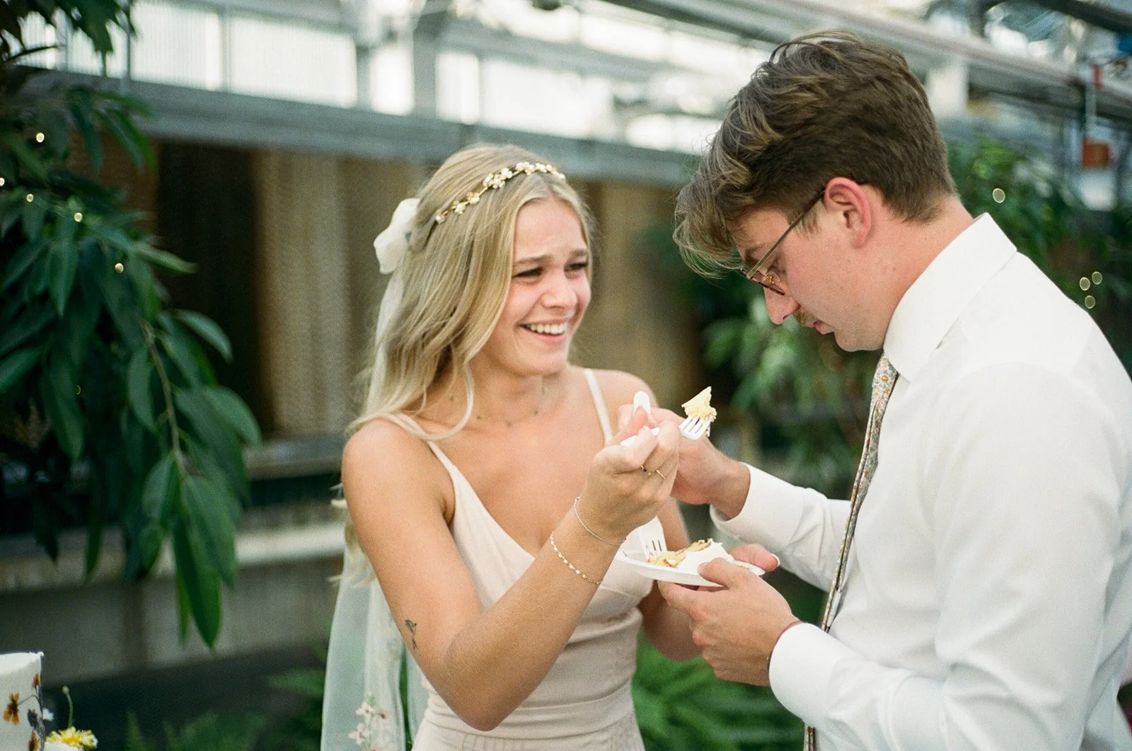 A woman with long blonde hair wearing a white dress and a floral headband is smiling as she feeds a man white cake with a fork. The man with glasses and brown hair is dressed in a white shirt and is holding a small plate and a fork.