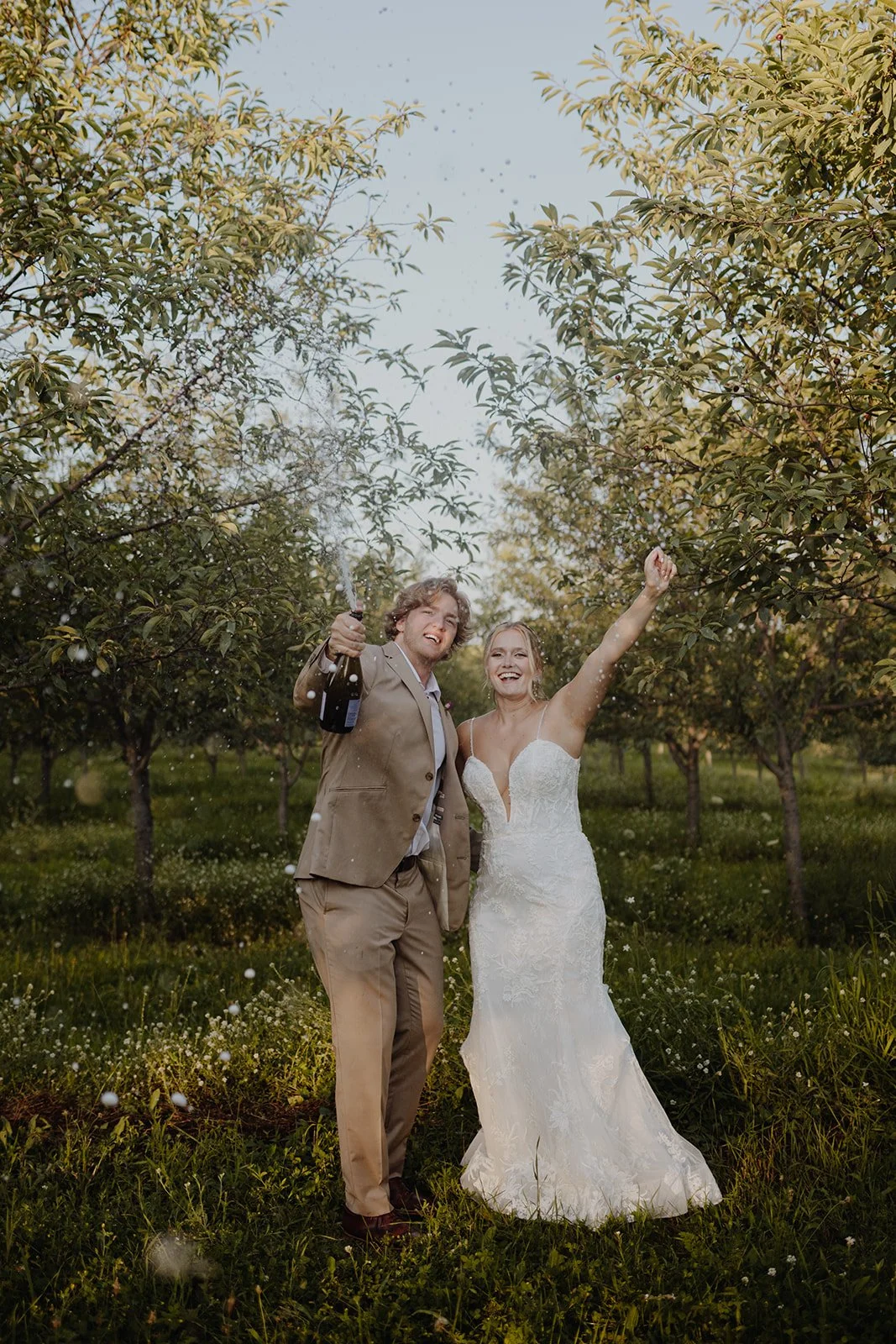 A newly married couple standing in a green orchard celebrating with champagne, smiling and raising their arms.