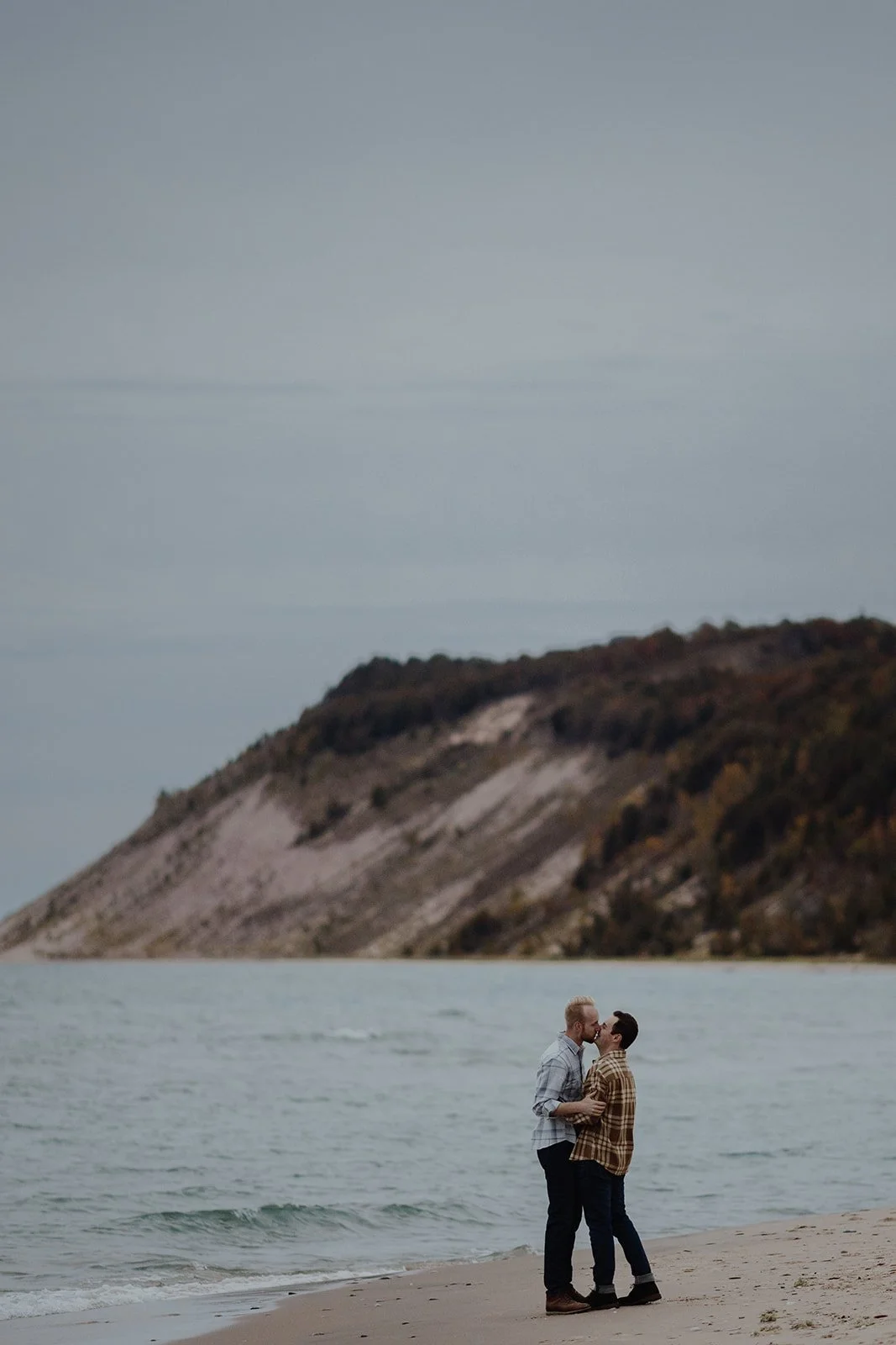 Two men kissing and holding each other on a beach with a large hill or mountain in the background.