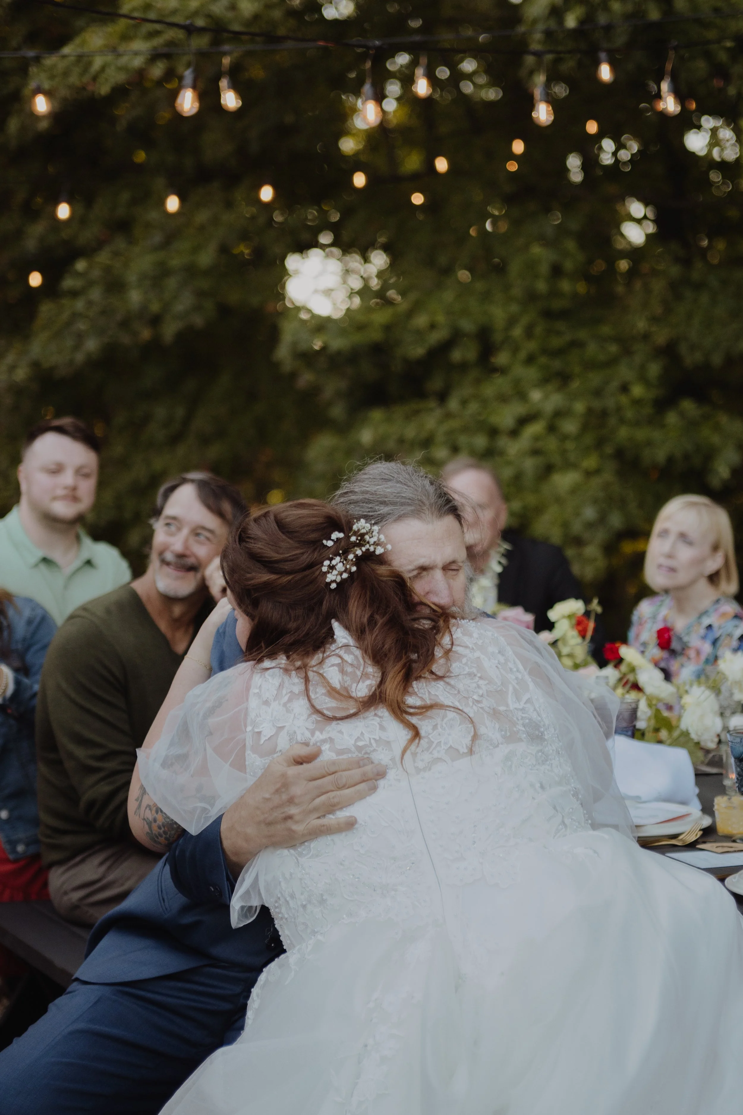 A bride and an older man, possibly her father, share an emotional hug at an outdoor wedding reception, with guests seated at a table decorated with flowers and string lights overhead.