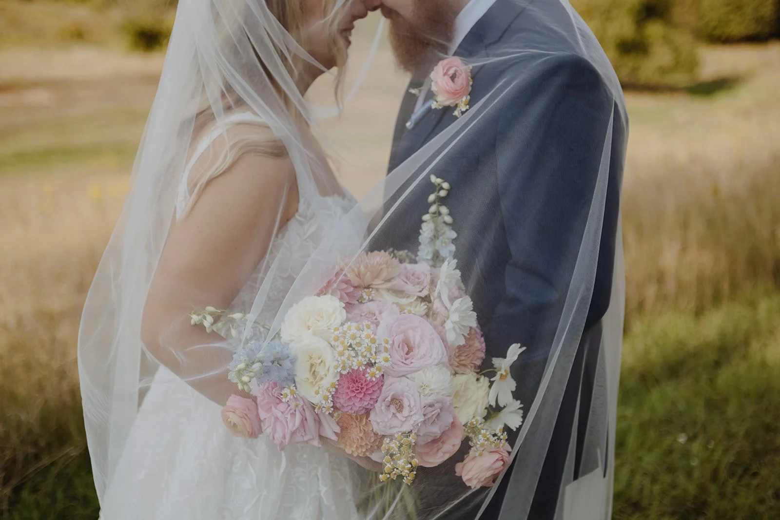 A bride and groom holding a bouquet of pink and white flowers, standing close together outdoors with a grassy field in the background.