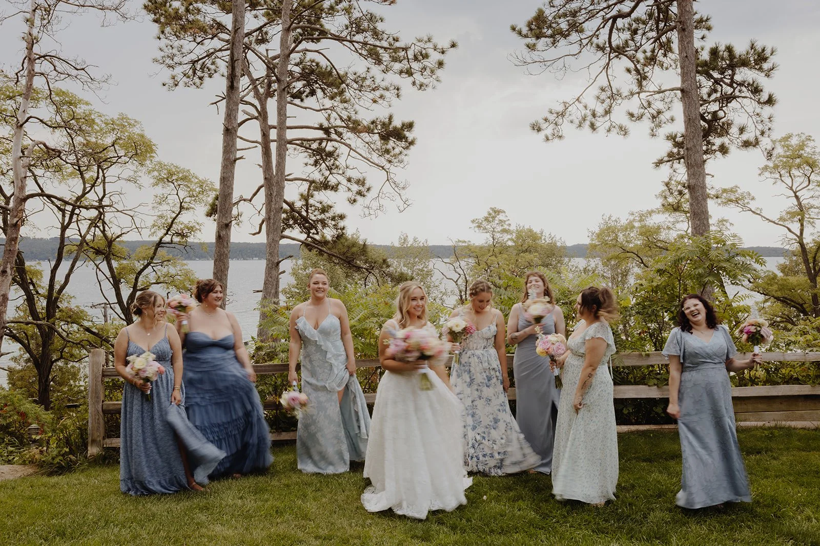 A group of women dressed in colorful dresses celebrating outdoors near a lake, holding bouquets of flowers, with trees and water in the background.