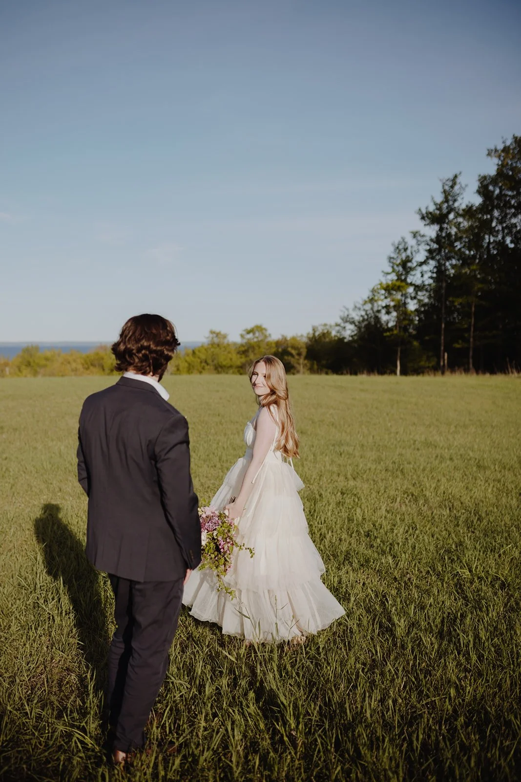 A couple standing in a grassy field, with the woman in a wedding dress holding a bouquet, and the man in a black suit, facing each other on a sunny day.