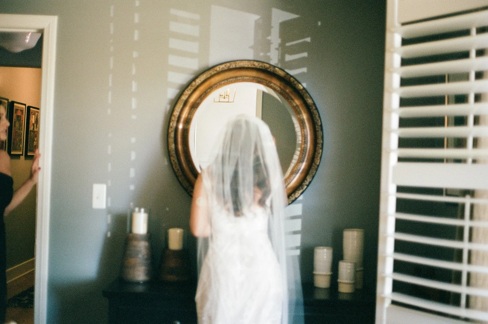 A woman in a wedding dress with a veil is looking into a large, ornate round mirror on the wall. An arm and part of a person's face are visible on the left side of the image, and there are candles on a table beneath the mirror.