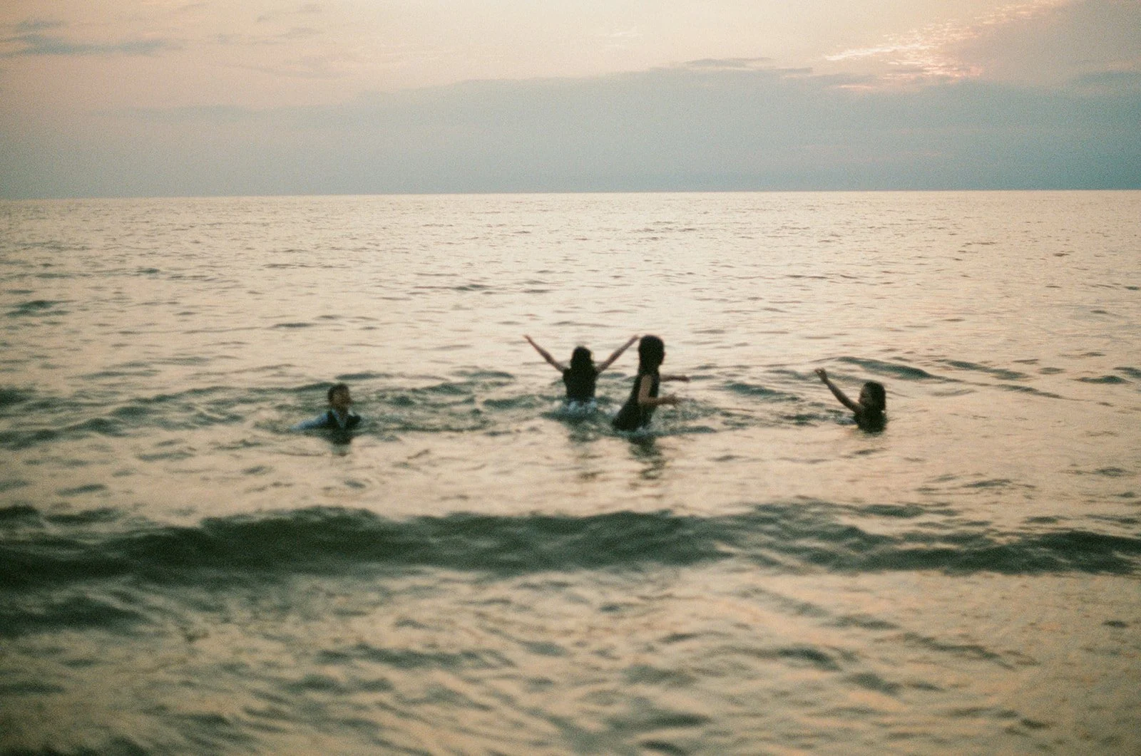 Four children playing in the ocean at sunset, with the sky showing soft clouds and the water reflecting warm light.