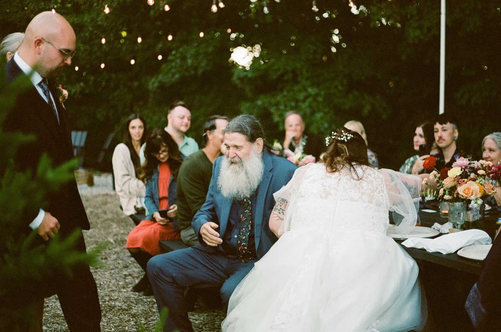 A wedding reception outdoors with a bride in a white gown and a groom surrounded by friends and family sitting at a table decorated with flowers, with string lights overhead.