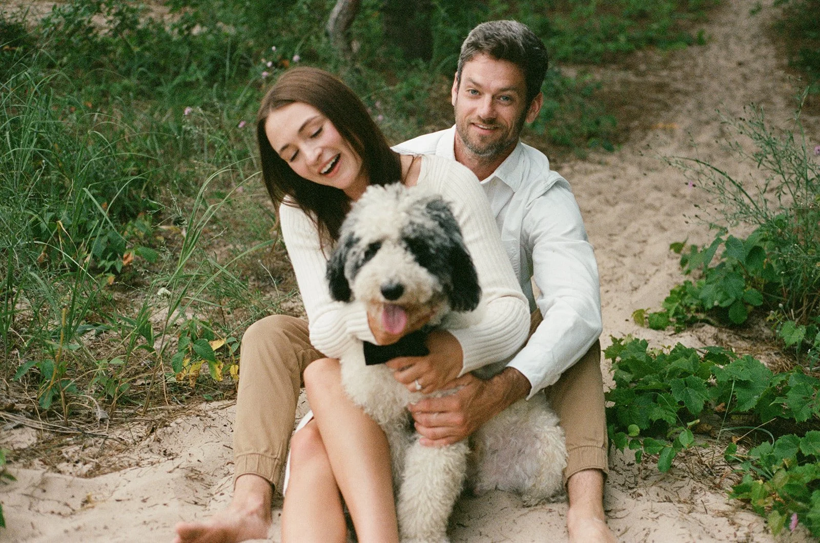A smiling woman and man sitting on the sand outdoors, holding a large fluffy black and white dog between them, surrounded by green plants.