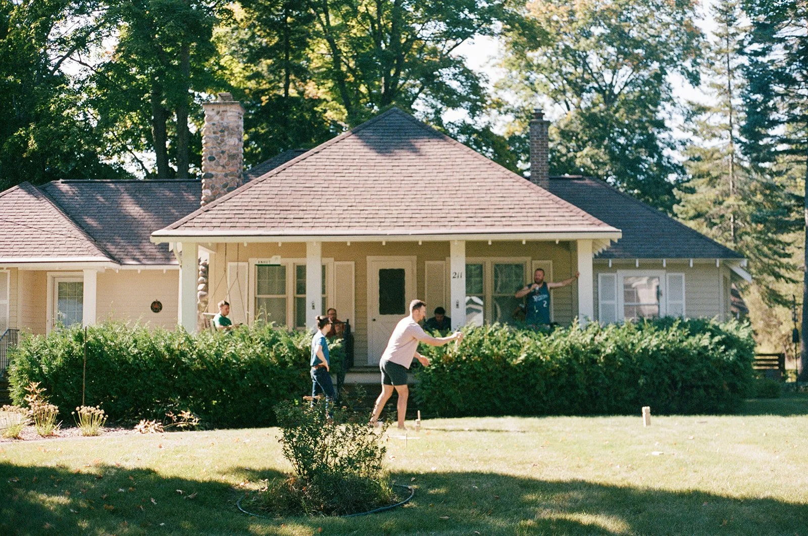 A group of people playing cornhole in the front yard of a house with a porch, surrounded by greenery and trees.