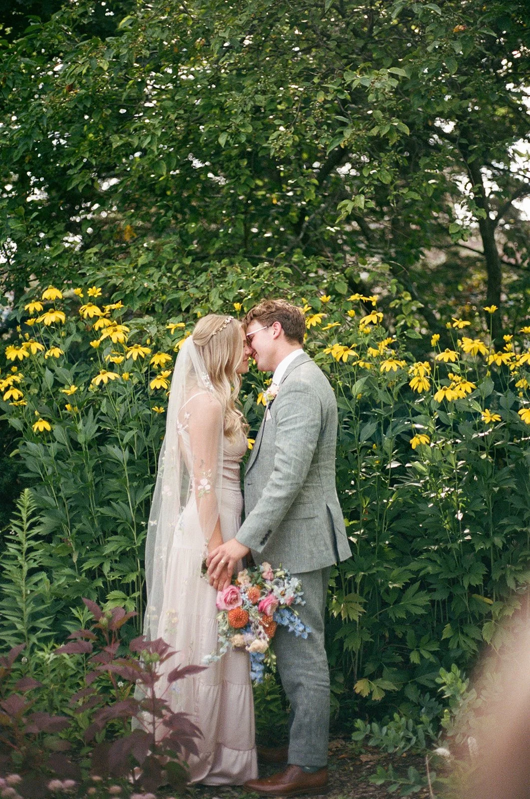 A bride and groom standing close with foreheads touching in a garden surrounded by yellow flowers and green foliage.