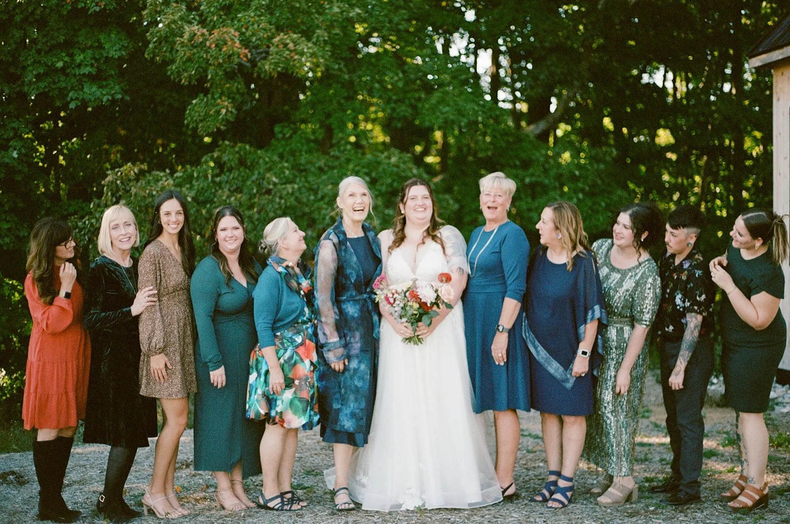 A group of thirteen women, including a bride holding a bouquet of flowers, standing outdoors in front of lush green trees, celebrating a wedding.