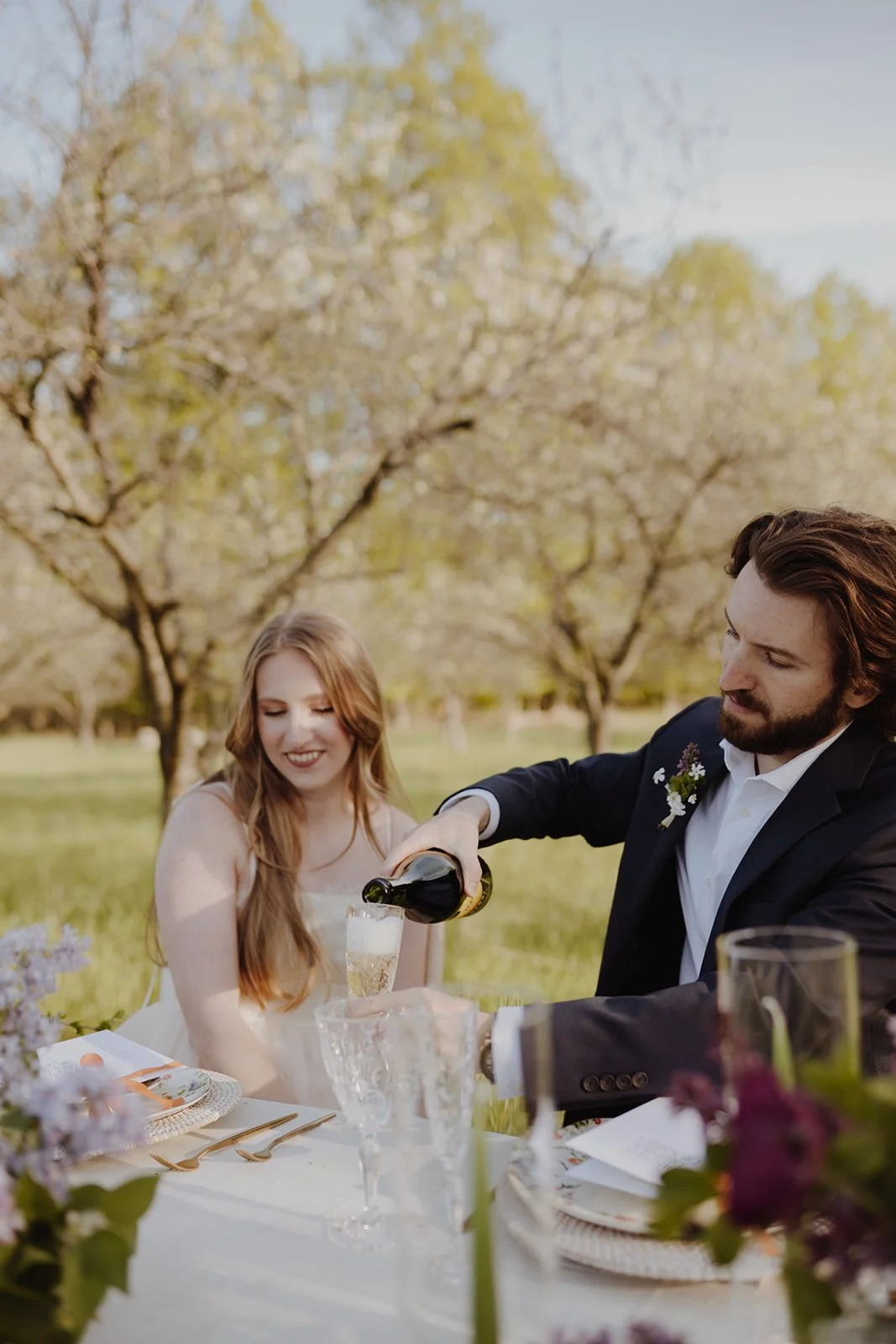 A man and a woman enjoy a picnic outdoors, with the man pouring a drink into a glass for the woman, in a park setting with trees and green grass.