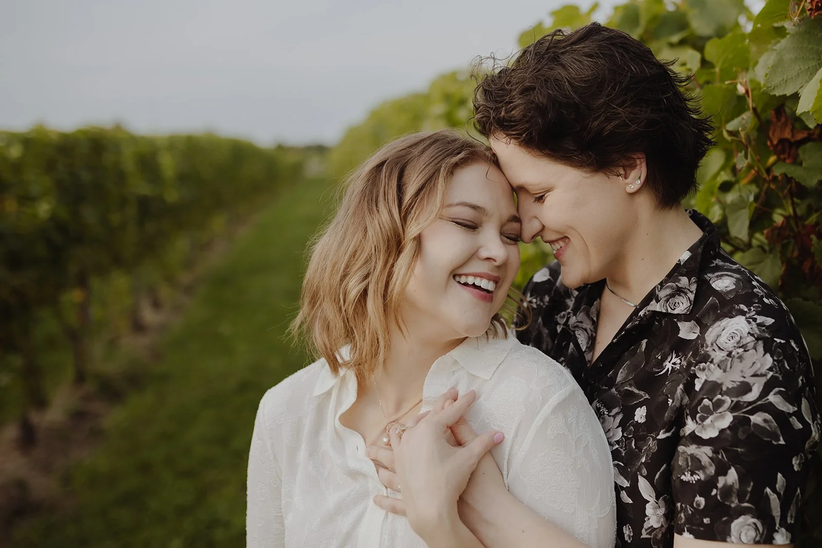 Two women share a joyful moment outdoors, smiling with their noses touching, in a vineyard setting.