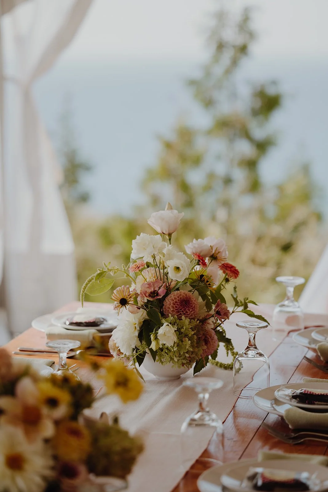 A floral centerpiece with pink, white, and green flowers on a dining table with wine glasses and plates, outdoors with trees and sky in the background.