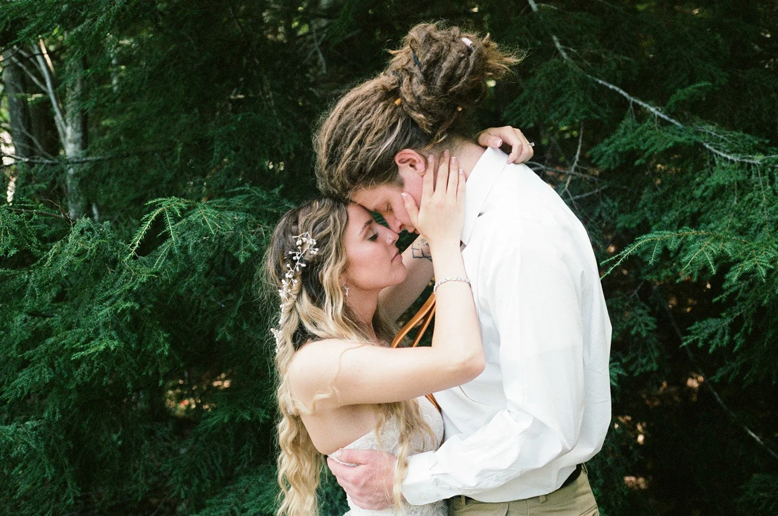 A couple is embracing closely outside, with the woman gently touching the man's face and both with their foreheads touching, surrounded by green foliage.