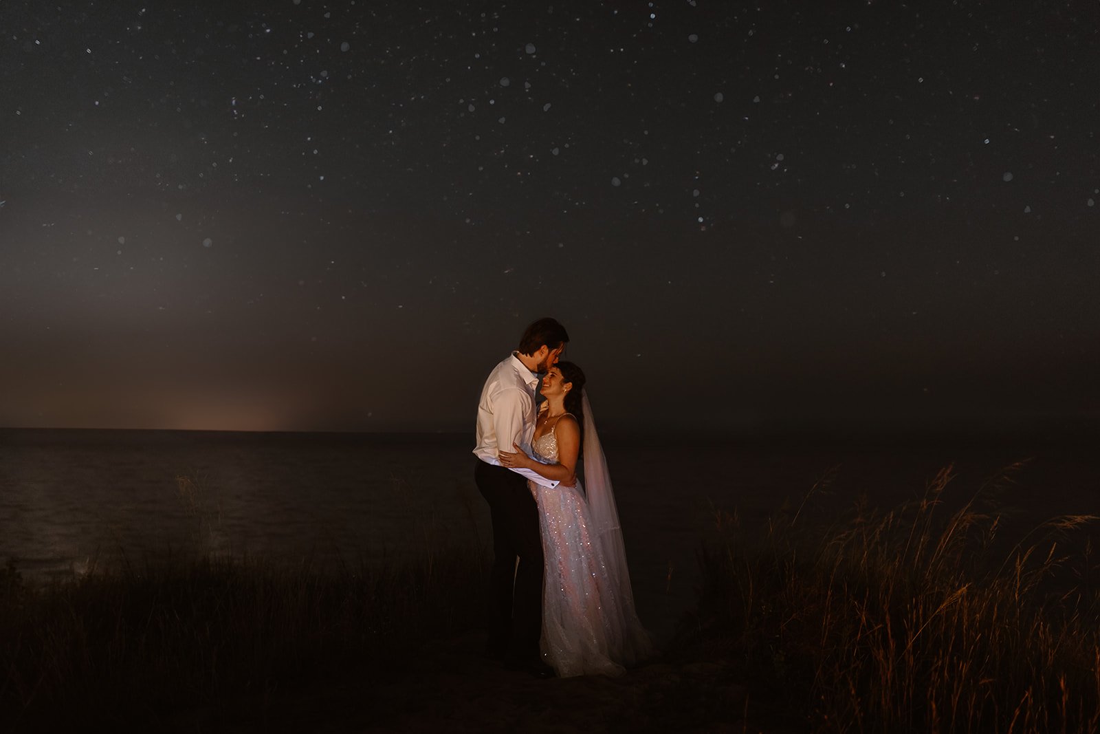 A couple in wedding attire embracing under a starry night sky by the water.