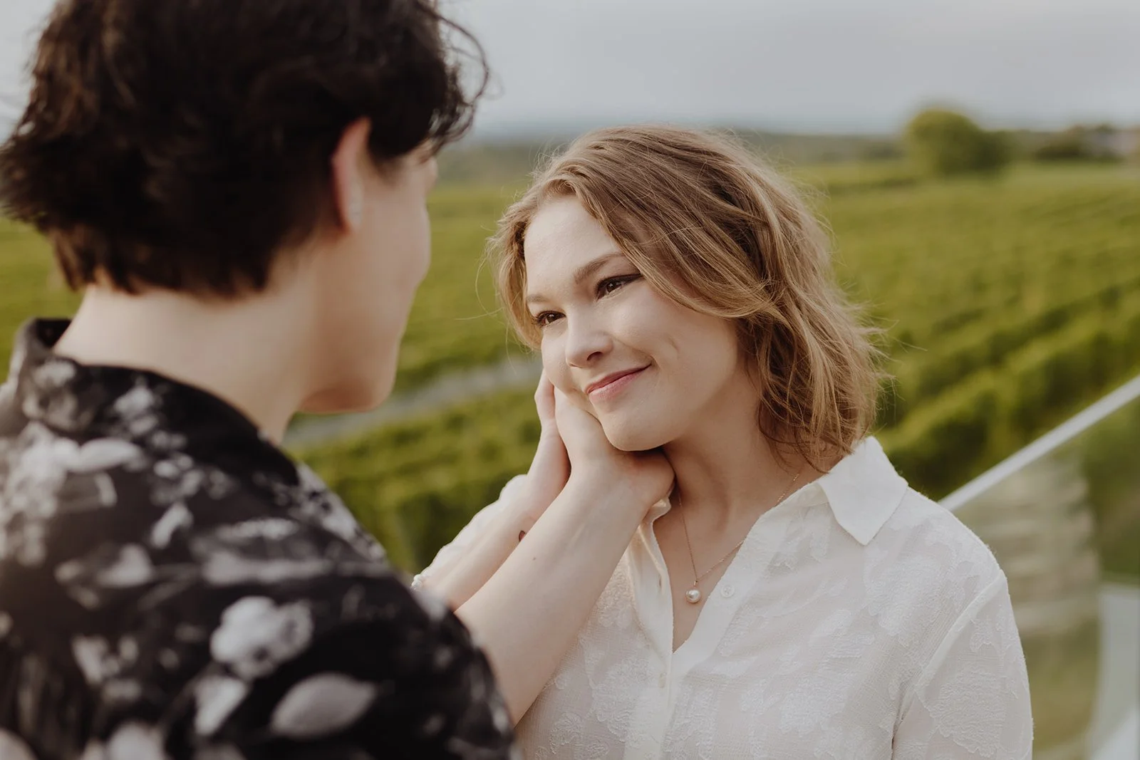 A woman with short, wavy reddish hair and a white blouse gently touching her cheek and looking at a man with dark curly hair and a black shirt with white floral pattern, outdoors in a green field or vineyard.