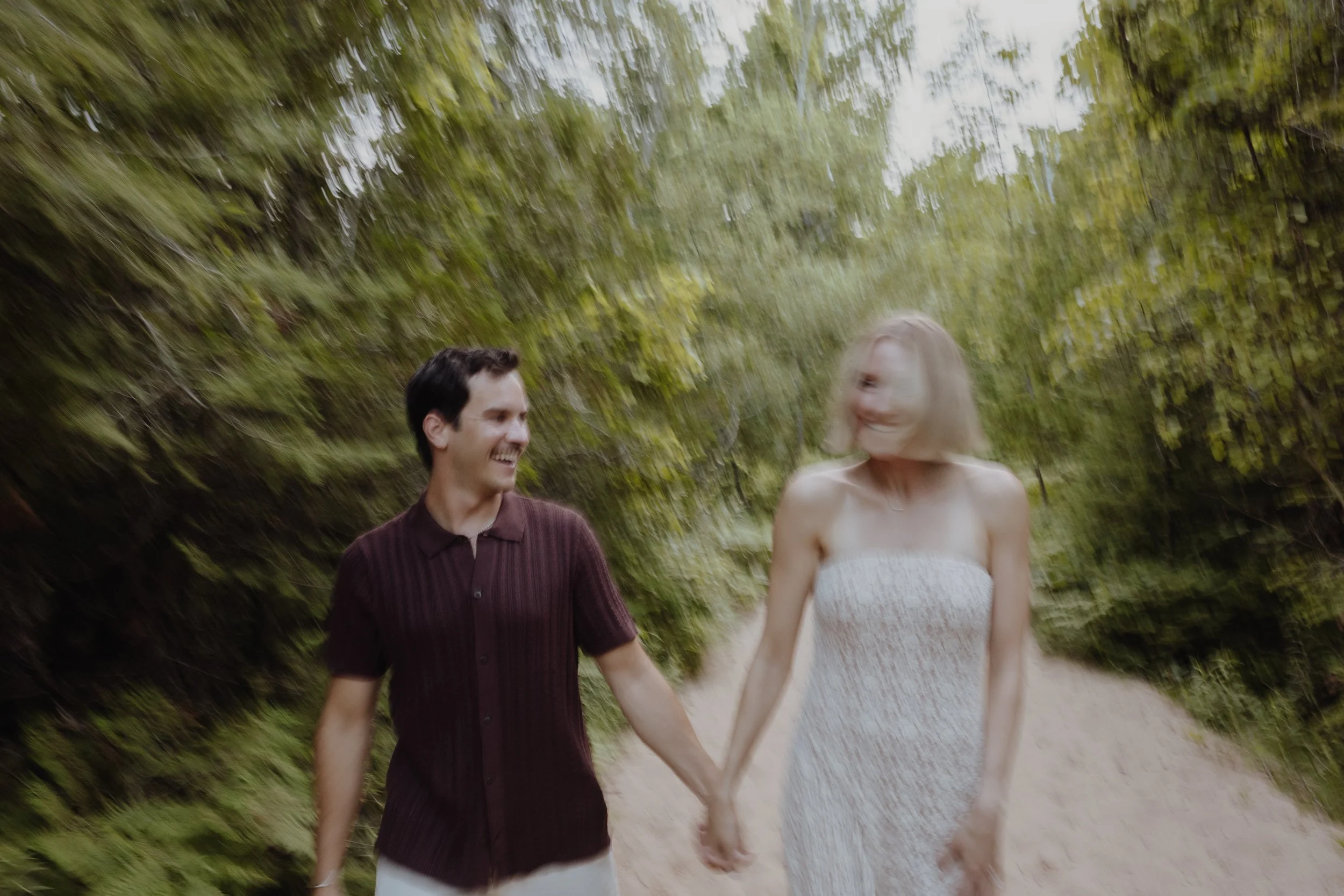 A smiling couple walks hand-in-hand through a wooded path, surrounded by green trees.